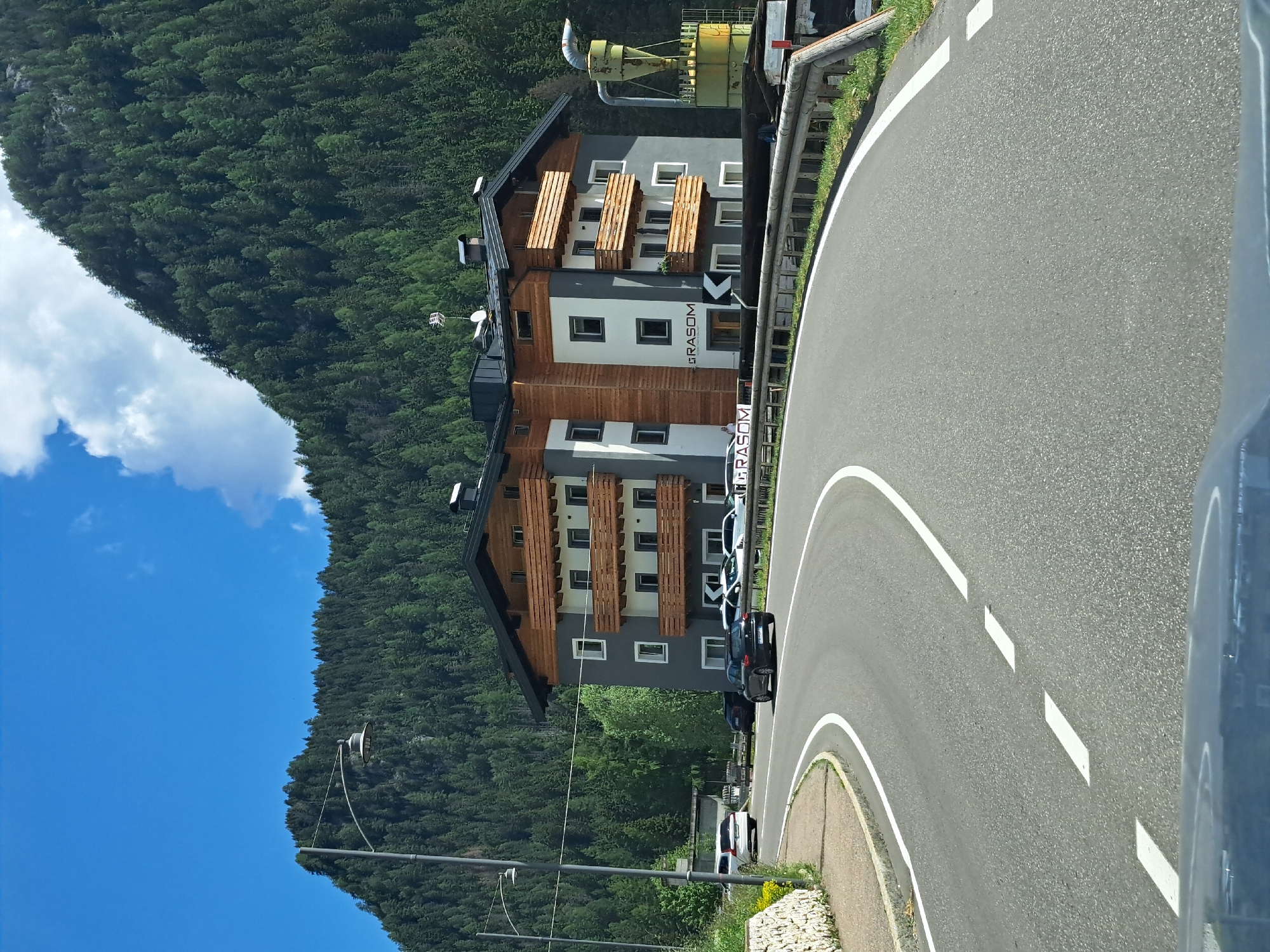 The image depicts a winding paved road curving to the right in a mountainous, forested area. On the left side of the road, a multi-story building with a modern design, featuring wood and grey facade elements, is situated against a backdrop of a dense green forest that covers a steep mountainside. Several cars are parked along the side of the building. The sky is a vibrant blue with scattered white clouds. The overall impression is one of a scenic, possibly tourist-oriented location, with the building likely being a hotel or similar accommodation. The time of day appears to be daytime, with clear weather. A large yellow industrial-looking structure with pipes is visible above the building. The road has white lane markings. Visible text on the building reads "GRASOM".