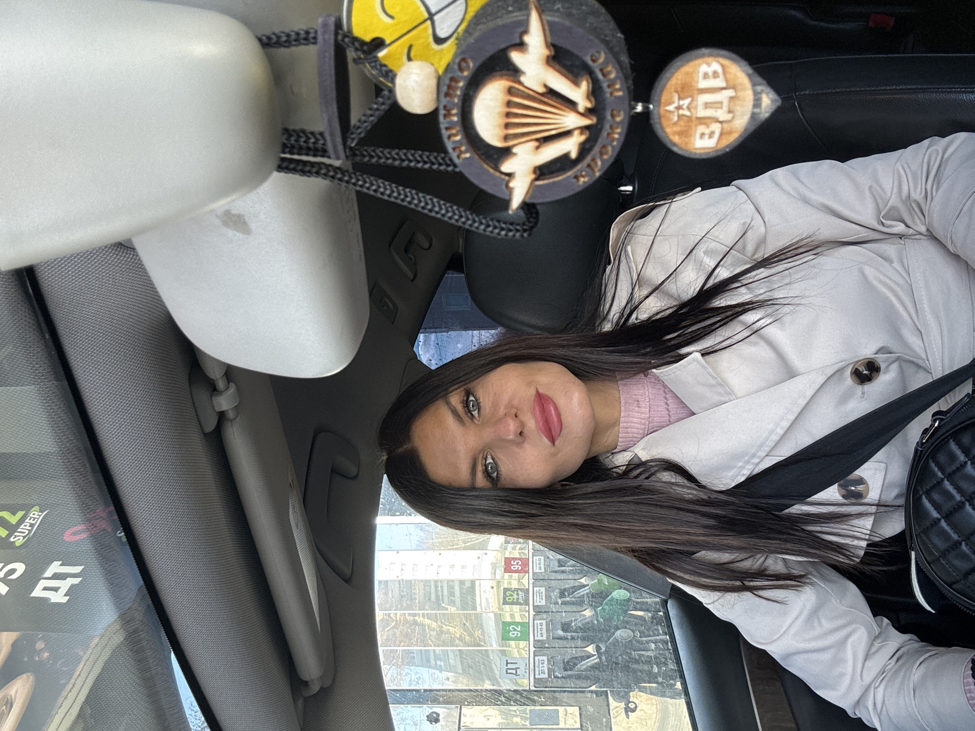 A woman with long brown hair is sitting in a car. She is wearing a light-colored trench coat over a pink sweater. The car is at a gas station, with fuel pumps visible through the window. The OCR text reveals that the gas station sells fuel types labeled as "92" and "SUPER". Car accessories, including a wooden decoration depicting paratroopers and another with the abbreviation "ВДВ," are hanging from the rearview mirror. The location is suggested to be in Chaikovsky, Russia.