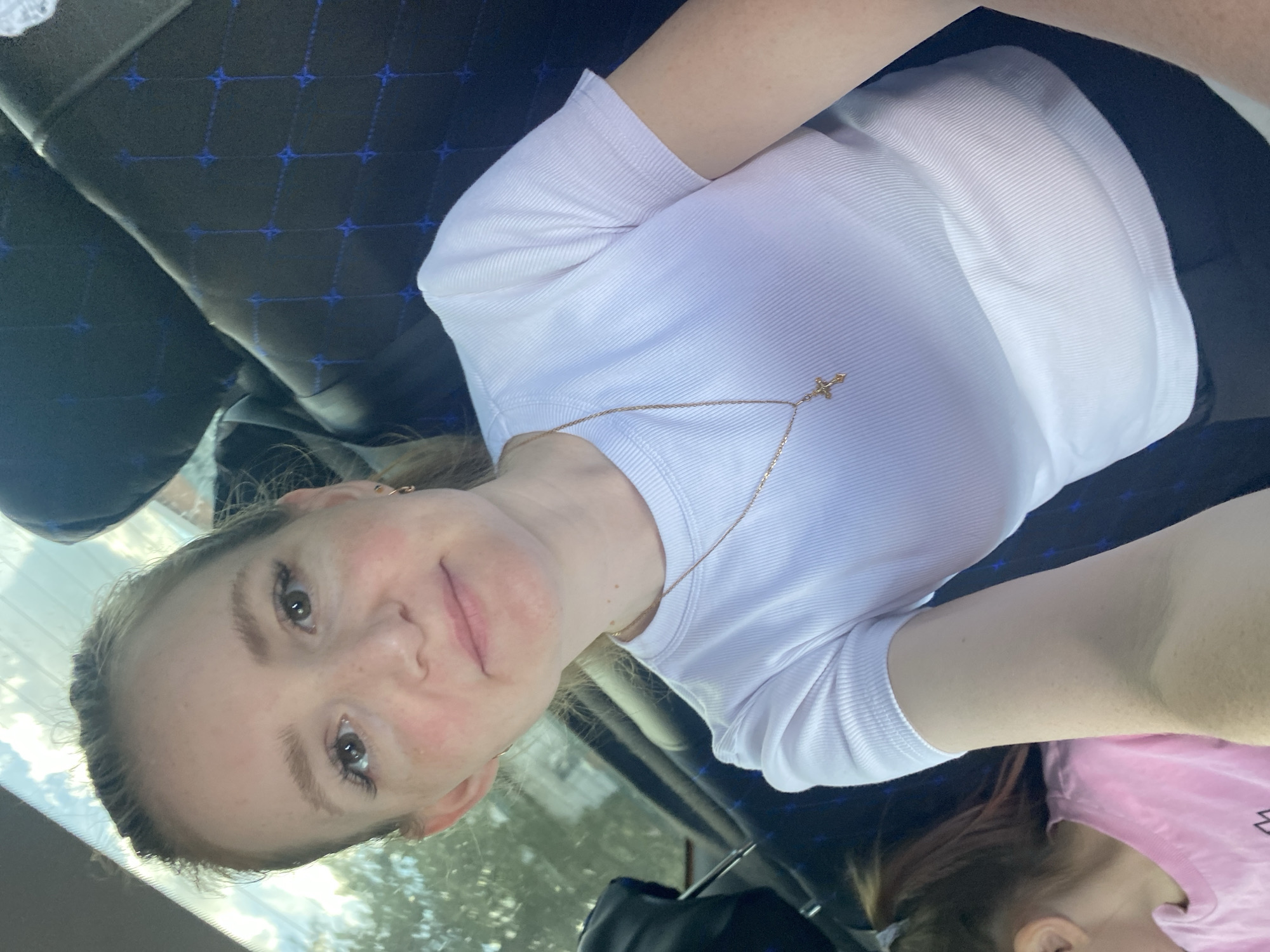 A woman with light skin and light brown hair pulled back is photographed from the chest up, looking into the camera. She is wearing a white, ribbed t-shirt and a gold necklace with a cross pendant. She is seated inside a vehicle, evidenced by the interior of seats with a blue diamond pattern visible behind her. Sunlight filters in from the front windshield, illuminating the scene. To her left, a portion of another person wearing a pink shirt is visible. The background suggests an outdoor environment with trees and sky visible through the windshield. The specific location in Chaikovsky, Russia, is not discernible from the visual elements presented.