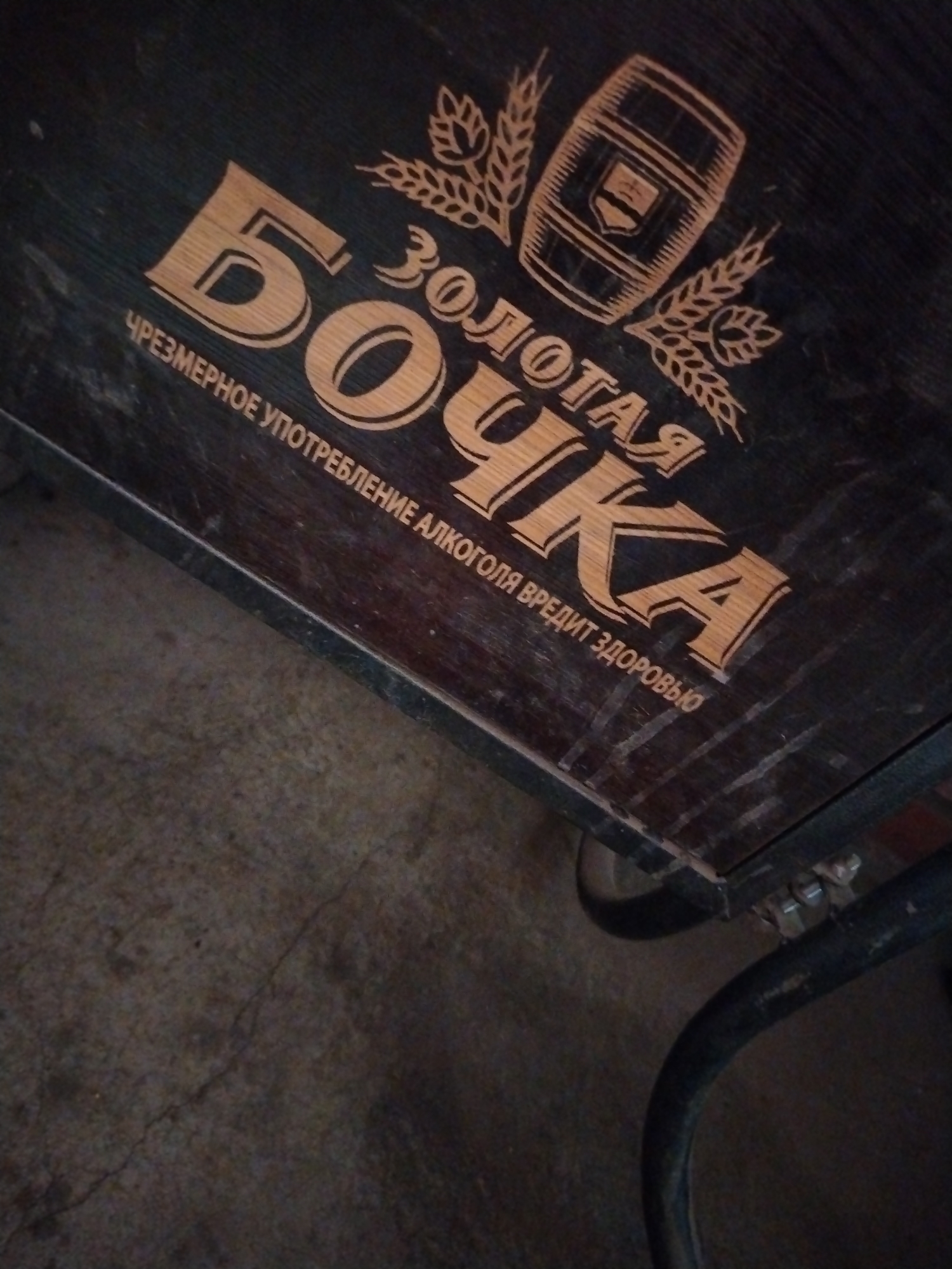 The image displays a close-up, slightly angled view of a dark wooden surface, likely the top of a cart or table, featuring an engraved or painted inscription in a lighter, wood-like color. The main inscription reads "ЗОЛОТАЯ БОЧКА" (Zolotaya Bochka), which translates to "Golden Barrel." This text is accompanied by an illustration of a wooden barrel flanked by wheat stalks. Below the main text, in smaller letters, there is a cautionary phrase: "ЧРЕЗМЕРНОЕ УПОТРЕБЛЕНИЕ АЛКОГОЛЯ ВРЕДИТ ЗДОРОВЬЮ," translating to "Excessive alcohol consumption is harmful to your health."
The wooden surface shows signs of wear, with some scratches and faded areas. In the lower right corner, parts of a dark metal frame and a wheel are visible, confirming that the object is a cart or a similar mobile structure. The ground beneath the object is a dark, uneven surface, appearing to be concrete or asphalt, with visible cracks and texture.
The lighting in the image is dim, casting a somewhat subdued and unembellished atmosphere. No people are present, and no specific activity is taking place; the image is a static shot focused on the inscribed object. The scene appears to be in a functional or outdoor setting in Шебунино, Russia.