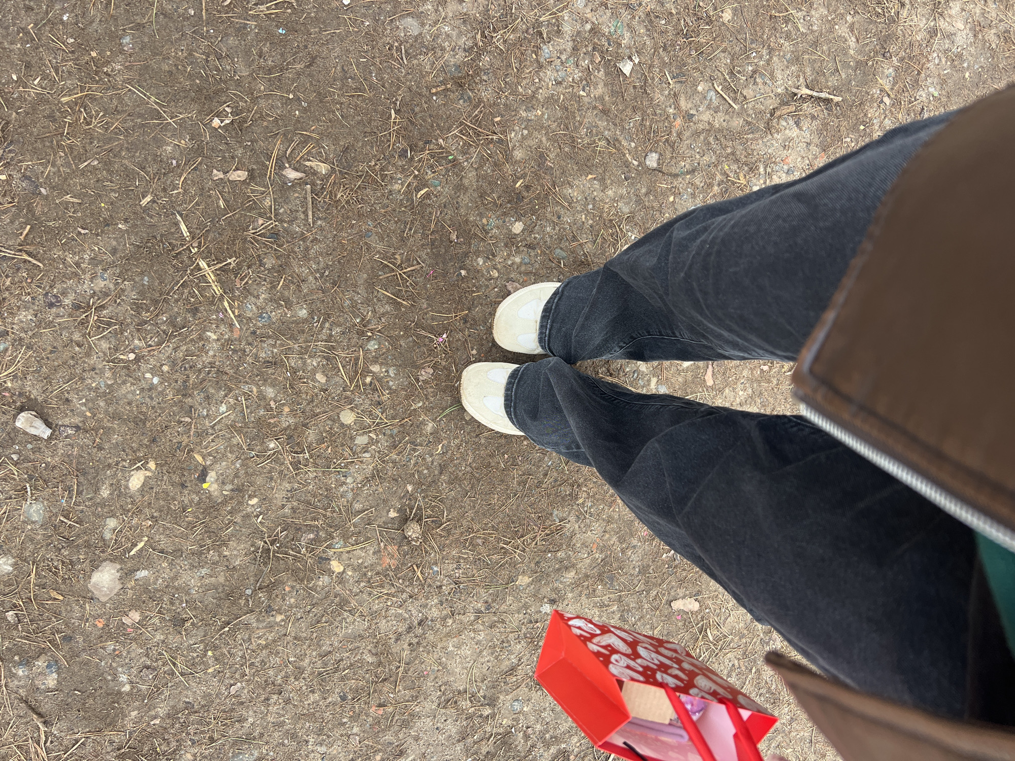 The image depicts a ground-level view of a person standing on a dirt and pine needle-strewn ground, with feet pointed slightly inward. The person is wearing dark-wash denim pants and off-white sneakers. A partially visible red gift bag with a white heart pattern is held in the lower right corner of the frame. The right side of the image is obscured by what appears to be a brown leather-bound item, possibly a bag or book.
Given the ground texture and sparse debris, the environment is likely an unpaved area, possibly a park or roadside. Without additional context, it is impossible to confirm the location in Chaikovsky, Russia.