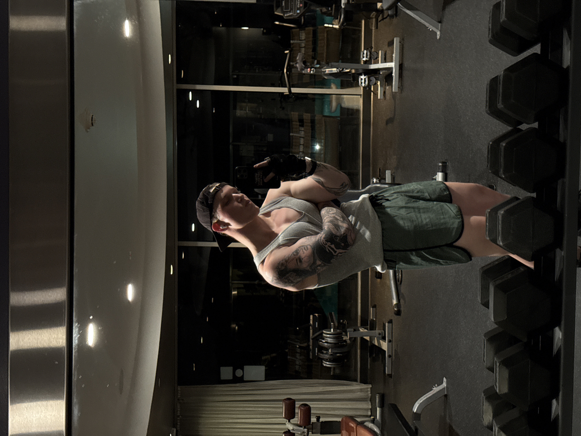 The image is a photograph taken in a gym, likely in West Hollywood, United States of America. The main subject is a muscular person wearing a tank top and shorts, with visible tattoos on their arms. They are holding a smartphone, presumably taking a selfie. The gym setting includes weightlifting equipment, such as a dumbbell rack with various weights, and other exercise machines. The environment appears dimly lit, possibly in the evening or early morning. The overall scene suggests a person documenting their fitness activity in a gym setting.