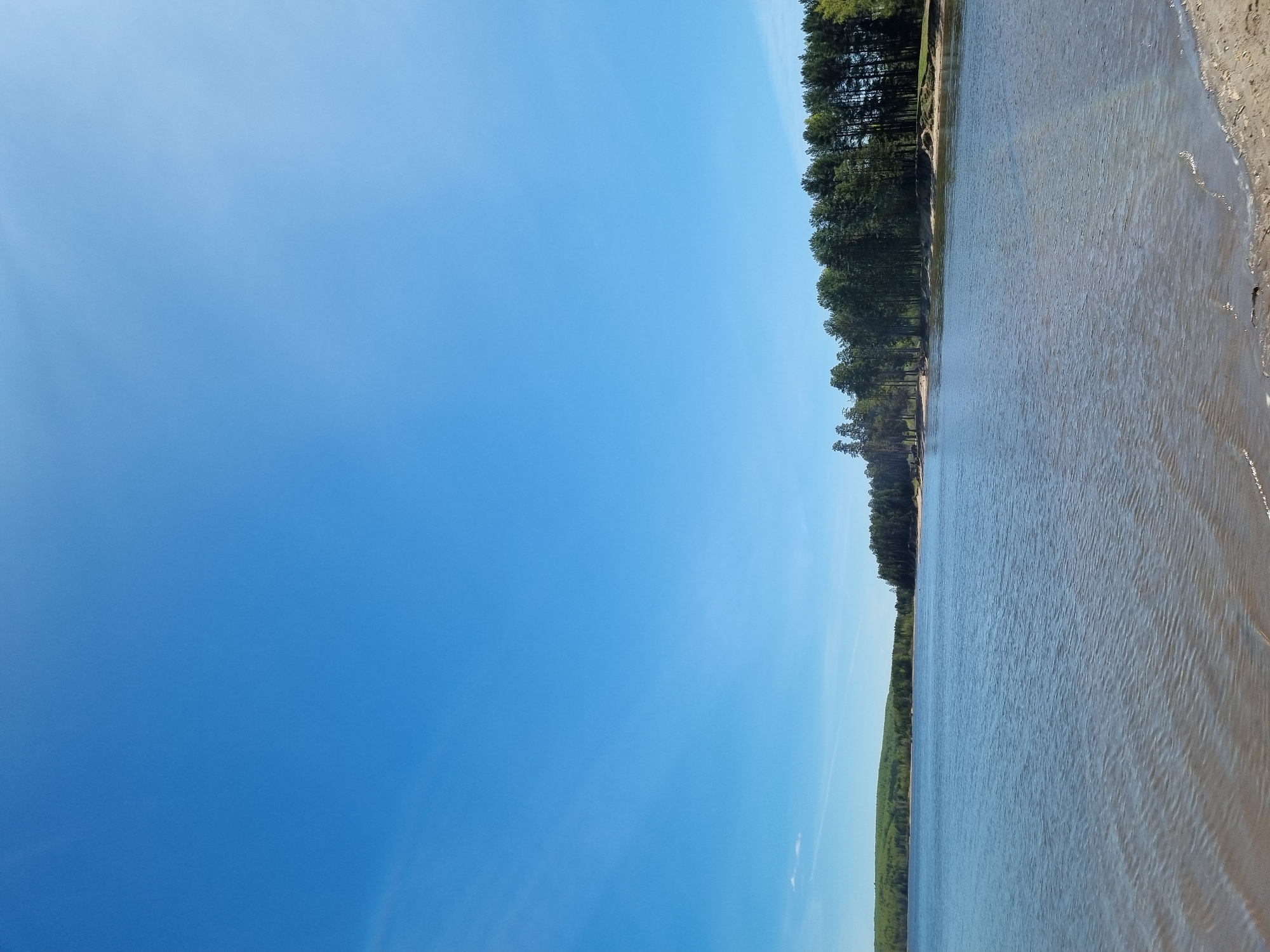 The image shows a landscape view of a body of water with a sandy shoreline on the right side. A line of trees is visible along the left side of the water. The sky, predominantly blue, occupies the upper portion of the image. The location is Chaikovsky, Russia.