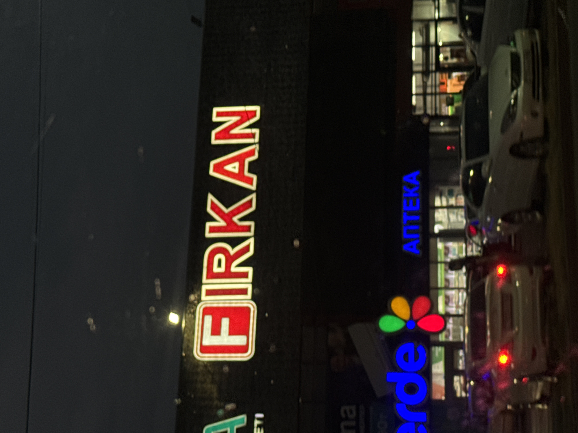 The image shows a nighttime street scene in Taraz city administration, Kazakhstan. A large, illuminated sign displays the word "FIRKAN" in red letters with white outlines, set against a dark background. Below this, another sign reads "VERDE" in blue letters, and to its right, "ANTEKA" in blue letters. The scene includes several cars, one white sedan is prominently positioned in the foreground. The overall environment appears to be an urban commercial area at night, with storefronts visible in the background.