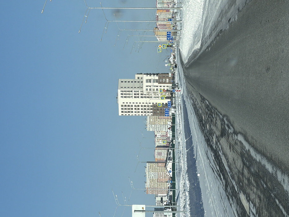 The image depicts a wide-angle, eye-level view of a snow-covered urban road in Tyumen, Russia, under a clear blue sky. The asphalt road, running from the foreground into the distance with a gentle curve to the right, is dark gray and partially clear, showing tire tracks and patches of melted snow and ice. Significant snowbanks line both shoulders of the road, indicating winter conditions.
Numerous streetlights with double-arm luminaires extend along both sides of the road. In the midground, a complex array of road signs is visible. These include multiple blue rectangular informational or directional signs, yellow diamond-shaped priority road signs, and yellow square signs with black pedestrian symbols indicating pedestrian crossings. Red and white striped traffic light poles are also present. A low green fence runs parallel to the road on the left side, behind the snowbank.
In the background, a modern, multi-story residential building complex, predominantly white with numerous windows and brown accents on its lower sections, dominates the right side of the frame. Beyond this, and extending across the horizon, are several other multi-story apartment buildings of varying designs and colors, including light brown, yellow, and beige facades, suggesting a mix of newer and older construction. Some smaller, individual houses are sparsely visible between the larger buildings. A few vehicles are discernible on the road in the distance, moving away from the viewer. No individuals are visible.