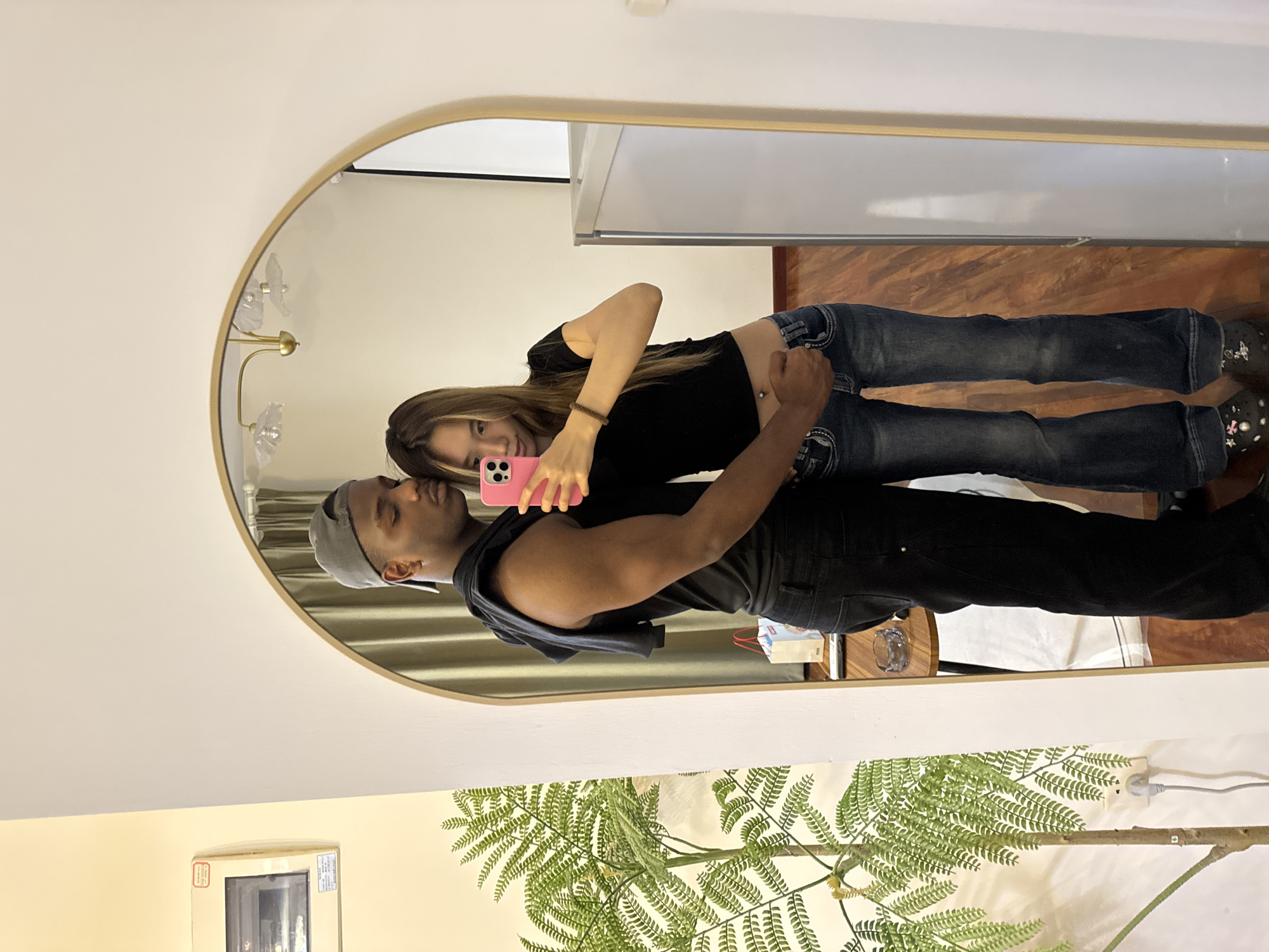 A reflection in an oval mirror shows a man and a woman. The man, wearing a gray baseball cap and a black tank top, stands behind the woman, who is wearing a black T-shirt and jeans. The woman holds a pink cell phone, presumably taking a selfie. The mirror also reflects part of a room. A white wall is visible, along with a window AC unit. In front of the wall is a faux fern plant. The reflections indicate that the space is in a residential setting in Yuzhong District, China. The mirror also reflects a light fixture.