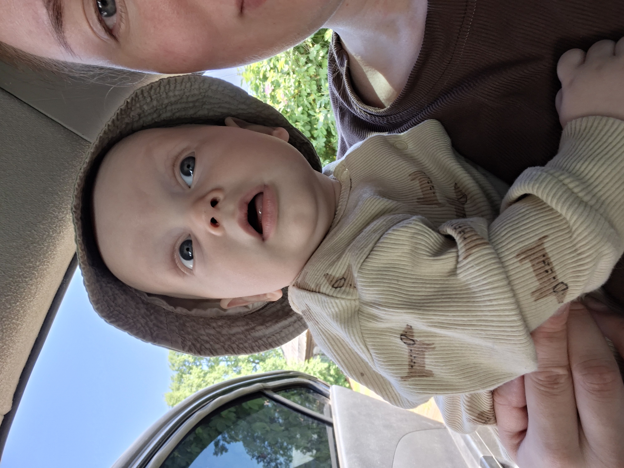 A close-up, canted shot shows a fair-skinned infant wearing a bucket hat and a striped shirt with brown animal designs. The baby is held by an adult with fair skin and a brown shirt. Part of a car interior and exterior are visible in the background, along with foliage and a blue sky. The location is not identifiable as Karsha, Russia, from the visual information provided.
