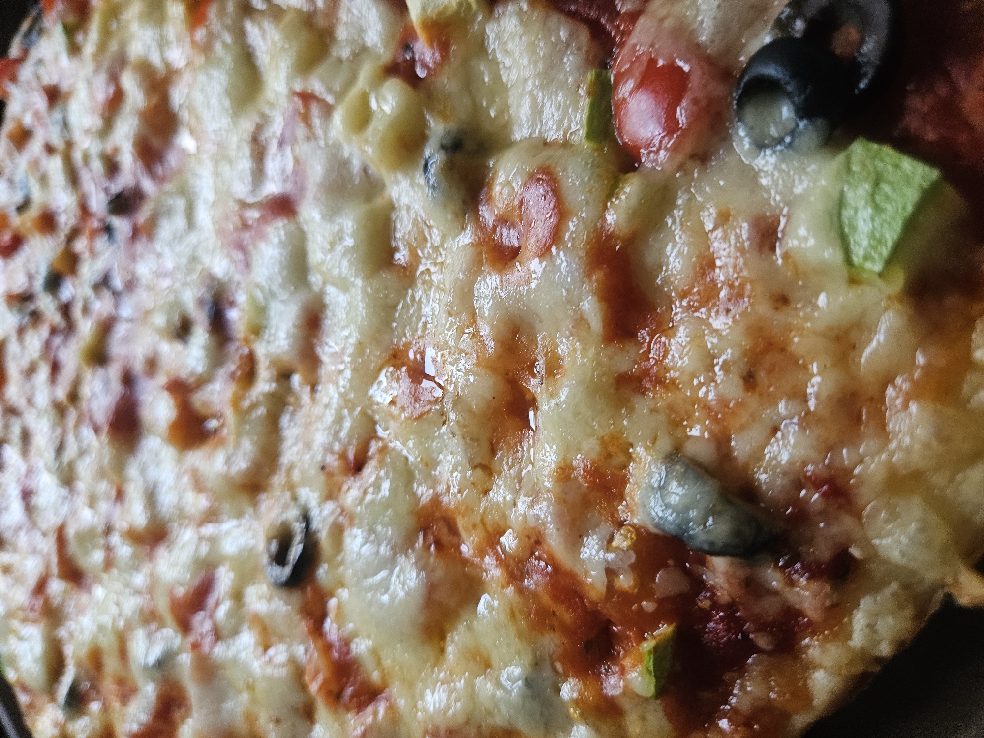The image is a close-up shot of a pizza, dominated by melted cheese. Toppings include what appear to be sliced olives, green peppers, and what may be pepperoni or diced tomatoes. The crust is visible along the bottom edge. There is no contextual information to suggest this image was taken in Rodniki, Russia, or to indicate any specific location, people, actions, or interactions.