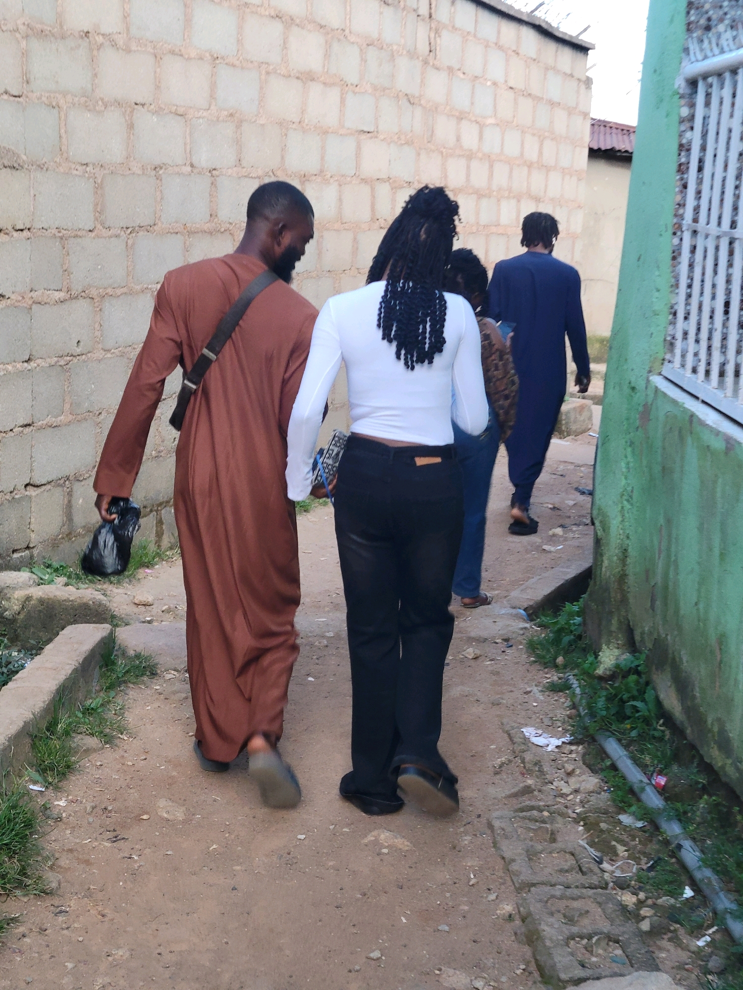 Four individuals, viewed from behind, walk along a narrow, unpaved path between two buildings in Jos, Nigeria. On the left, a man wearing a long, brown robe and a dark strap across his shoulder carries a black plastic bag in his right hand. Beside him, to his right, a person in a white long-sleeved shirt and black pants walks with a patterned clutch bag. Behind them, a second person wearing a dark blue robe is visible. The path is bordered by a textured, light-colored brick wall on the left and a green wall on the right. Debris, including stones and scattered white paper, is present on the ground. The environment appears to be an urban, possibly residential area, with a rough, utilitarian aesthetic.
