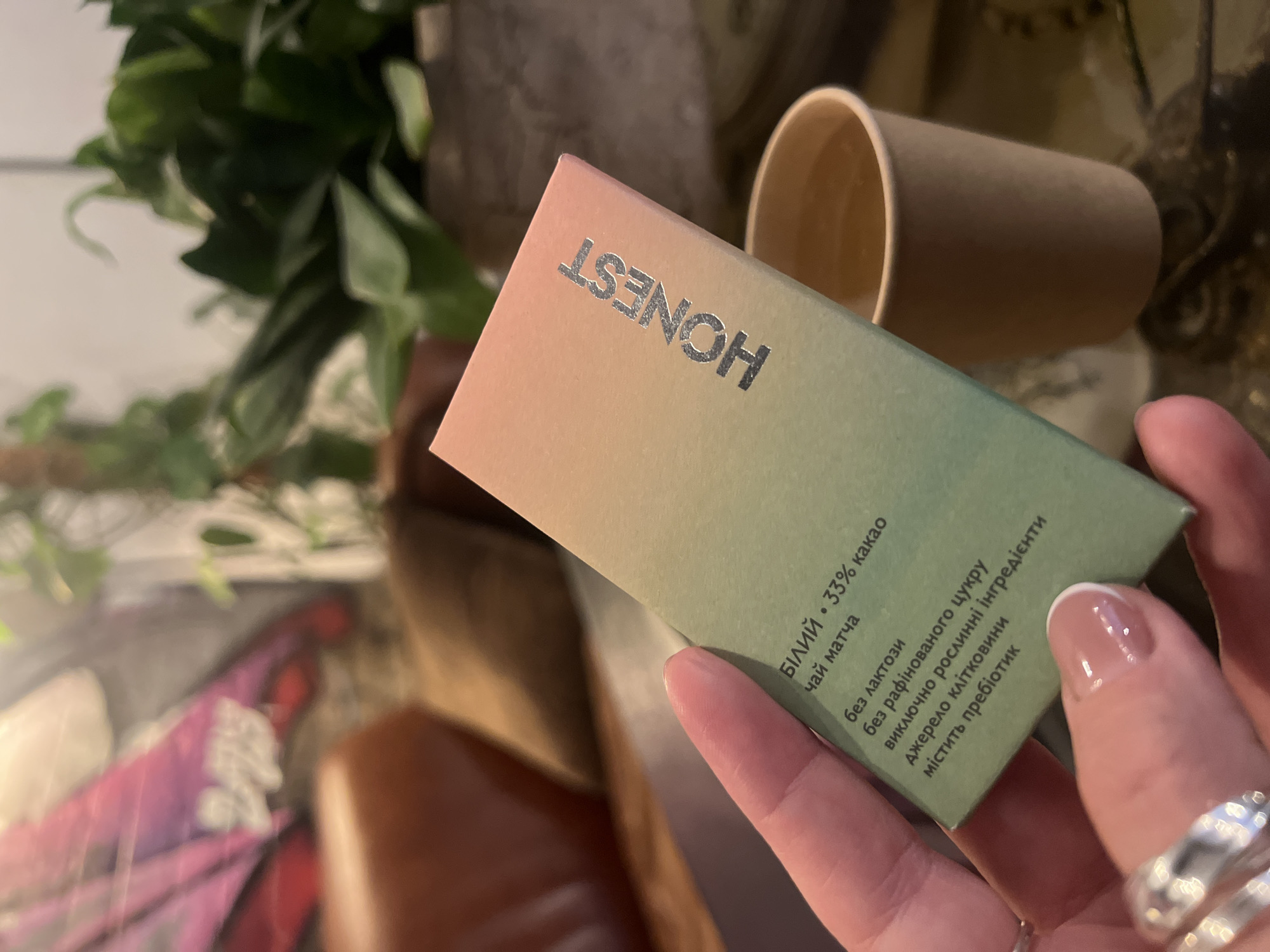 A close-up shot captures a person's hand holding a small, rectangular package of Honest brand chocolate. The package is a gradient of soft pink, orange, and green, with the word "HONEST" embossed in silver foil. Text on the green side of the package is in Ukrainian and indicates the chocolate is white with 33% cacao, matcha tea, lactose-free, without refined sugar, exclusively from plant-based ingredients, and contains prebiotics. A woman's fingers, adorned with rings on two fingers, are visible holding the package. In the background, out of focus, is a tan-colored disposable cup containing a light-colored liquid, and a blurred potted plant with green leaves. The background also shows hints of indoor furniture and lighting, suggesting a cafe or similar establishment. There is no visual information within the image to suggest a specific location in Kyiv, Ukraine.