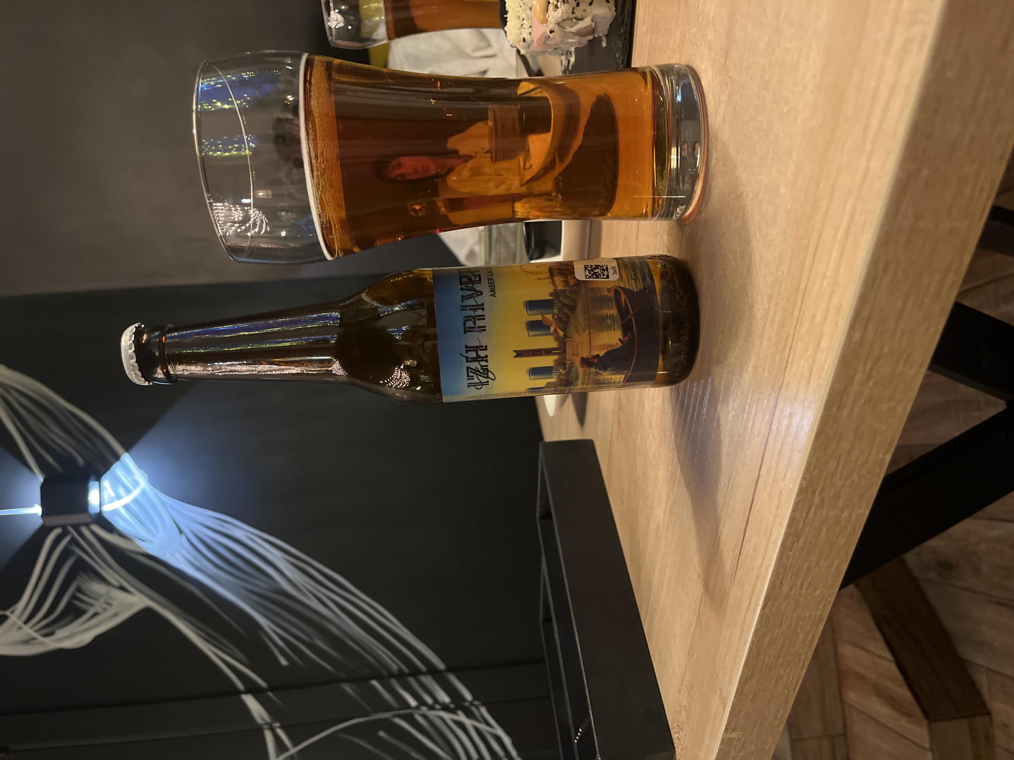 This media file captures an indoor scene, likely in a restaurant, bar, or pub, featuring a glass of beer and a beer bottle on a wooden table. The lighting suggests evening or nighttime.
The main subjects are a half-full glass of amber-colored beer with a thin head, and a brown glass beer bottle standing next to it. The beer appears to be an amber lager. To the right of the glass, partially out of frame, is a small dish containing what appears to be sushi or similar small bites.
A notable detail is the reflection of a person's face and upper body visible in the beer glass. The person has short, dark hair and appears to be looking towards the camera's position. The reflection also shows glimpses of the surrounding interior, possibly including other tables or chairs.
The beer bottle features a label with a light blue and yellow design depicting a landscape, possibly a river and buildings. Visible text on the label includes "IZH RIVE" and "AMBER LAGE" (likely Amber Lager). This strongly suggests a connection to the Izhevsk River, aligning with the provided location context of Izhevsk, Russia. A QR code is also present on the bottle label.
The background consists of a dark wall decorated with bright, abstract, flowing lines, illuminated by a focused light source from the left side of the frame, possibly creating a modern or artistic ambiance. The activity implied is enjoying a drink and possibly a meal in a relaxed setting.
