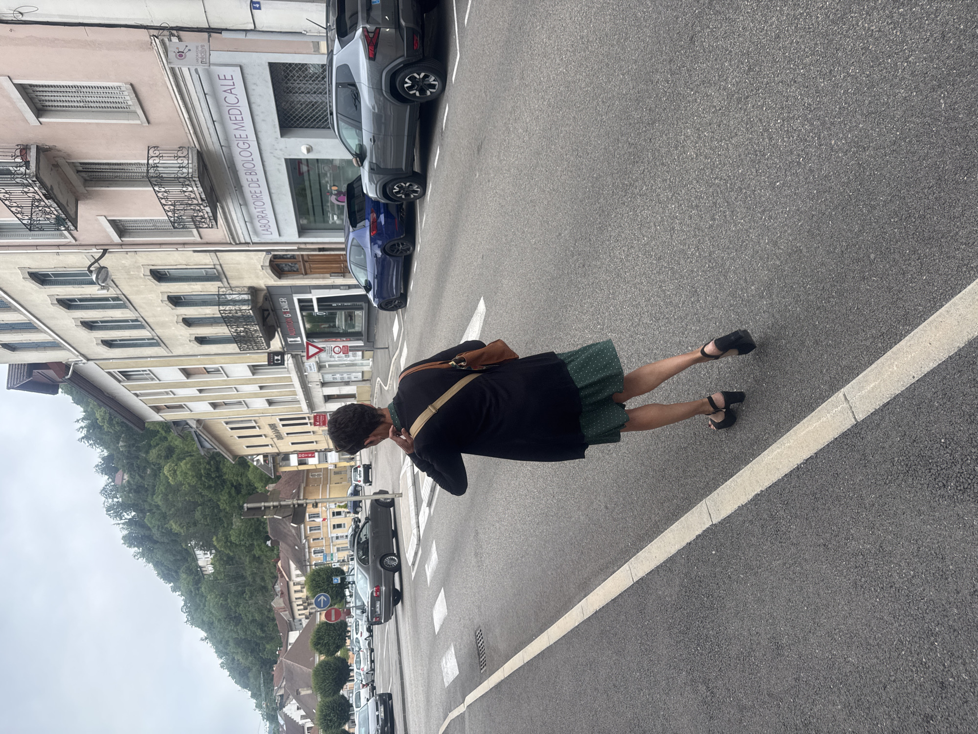 The image shows a street scene in Saint-Claude, France. In the foreground, a person wearing a green polka-dot dress and a black cardigan is walking away from the camera, facing towards a crosswalk and intersection. They are carrying a brown crossbody bag. In the background, there are buildings lining the street, a hillside covered in trees and some houses, and several parked and moving cars. One building prominently displays a sign that reads "LABORATOIRE DE BIOLOGIE MEDICALE." The sky is overcast.