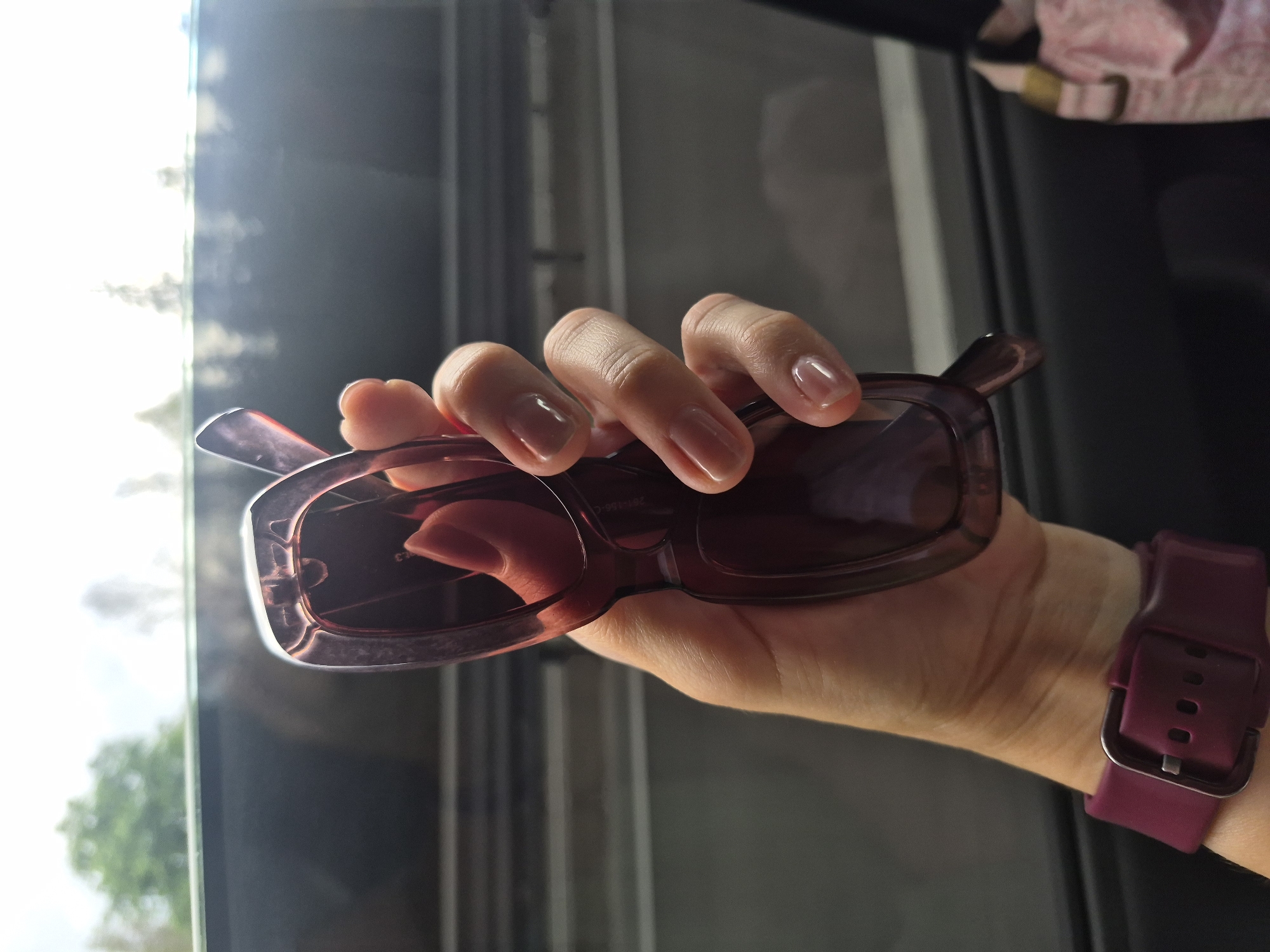 A close-up shot shows a person's left hand holding a pair of dark, translucent sunglasses. The hand has short, clear-polished fingernails and is positioned in front of a blurred background that appears to be a view from inside a vehicle looking out a window. The outer part of the sunglasses frame is visible, with one lens partially obscuring the hand. A dark purple watch strap is visible on the person's wrist. The environment suggests movement, possibly traveling by car or train, with horizontal lines of what could be seats or a structure visible in the middle ground. The lighting is bright and diffused, indicating it might be daytime with overcast conditions or the sun is behind the viewer. The image is sharpest on the hand and sunglasses. There are no discernible features that would indicate the location is specifically in Старые Решёты, Russia, as the image lacks identifying landmarks or context.