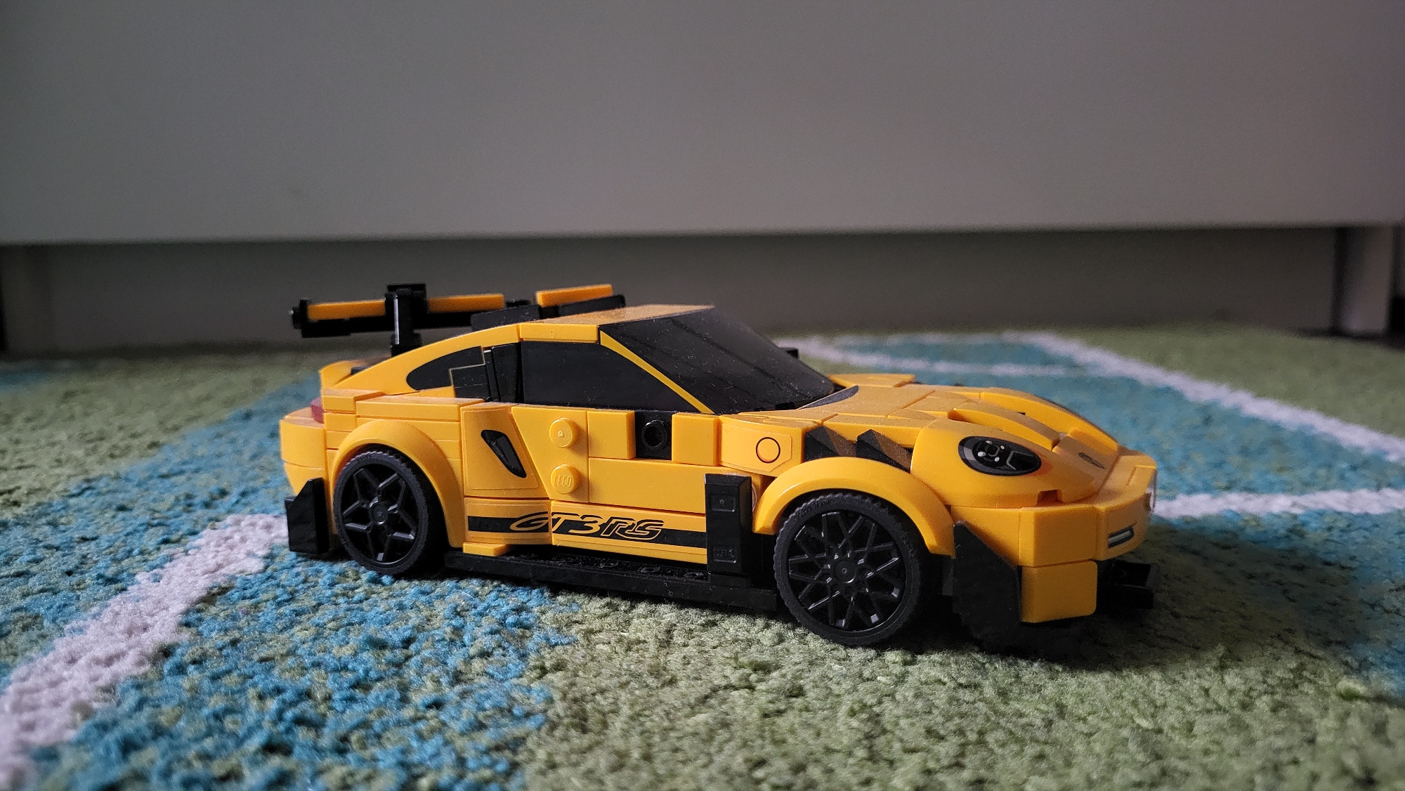 A yellow Lego Porsche 911 GT3 RS model car is on a carpet with a blue and green pattern and white stripes. The car is oriented at an angle to the viewer, showing the side and front. The car has black wheels and a rear spoiler. Behind the car is a white wall. The location is not identifiable as Sofia, Bulgaria.