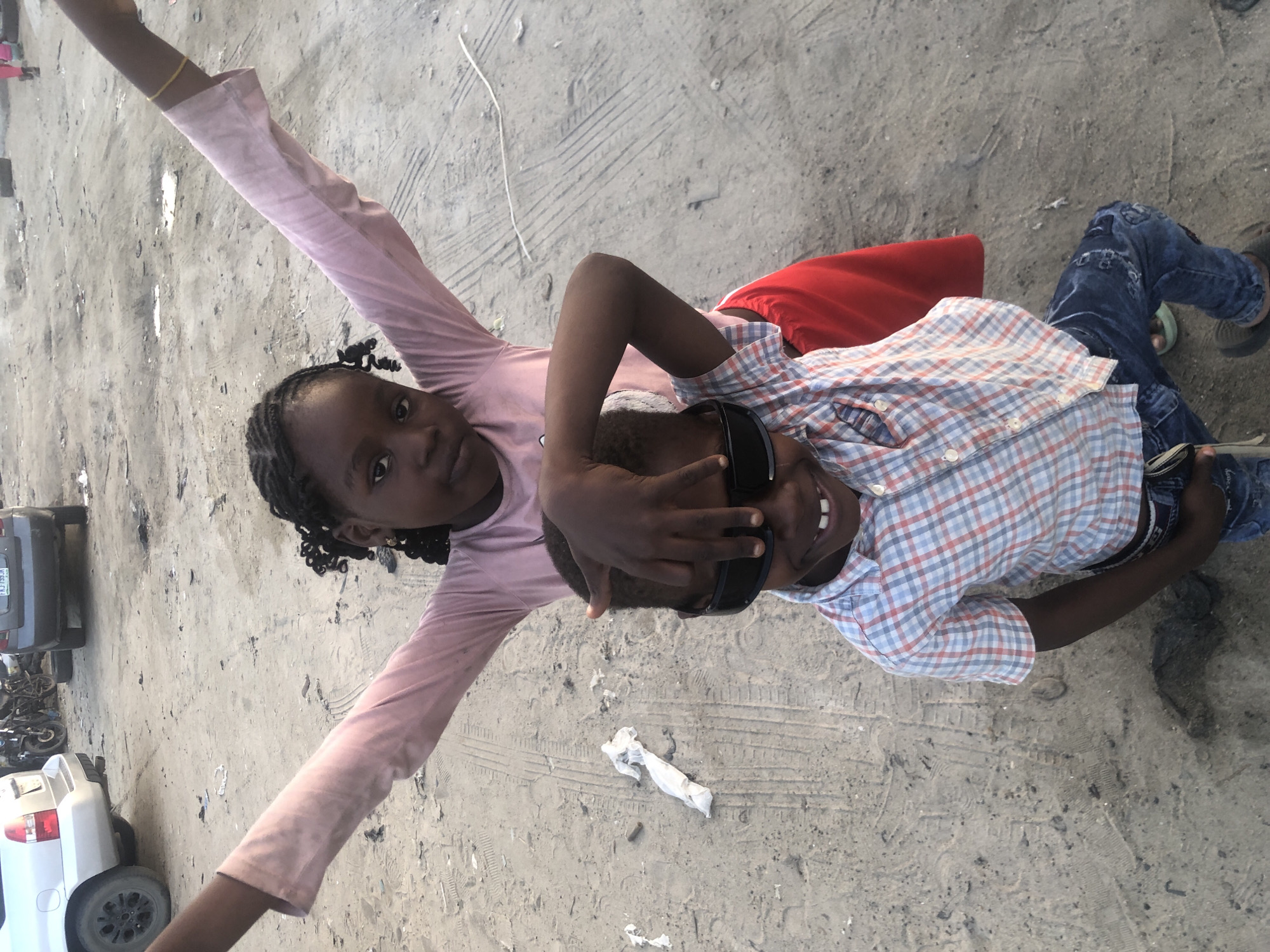 In Ado, Nigeria, two young children are pictured on a dirt ground. The older child, a girl, stands behind the younger child, a boy, with her arms outstretched, wearing a pink long-sleeved shirt. Her face is turned towards the camera, and her hair is styled in braids. The boy in front is wearing a plaid shirt, jeans, and dark sunglasses, with his hand resting on his head, partially obscuring his face. He is smiling, revealing his teeth. The background shows a rough, sandy area with tire tracks, scattered debris, and a few vehicles parked in the distance, suggesting an urban or semi-urban setting.