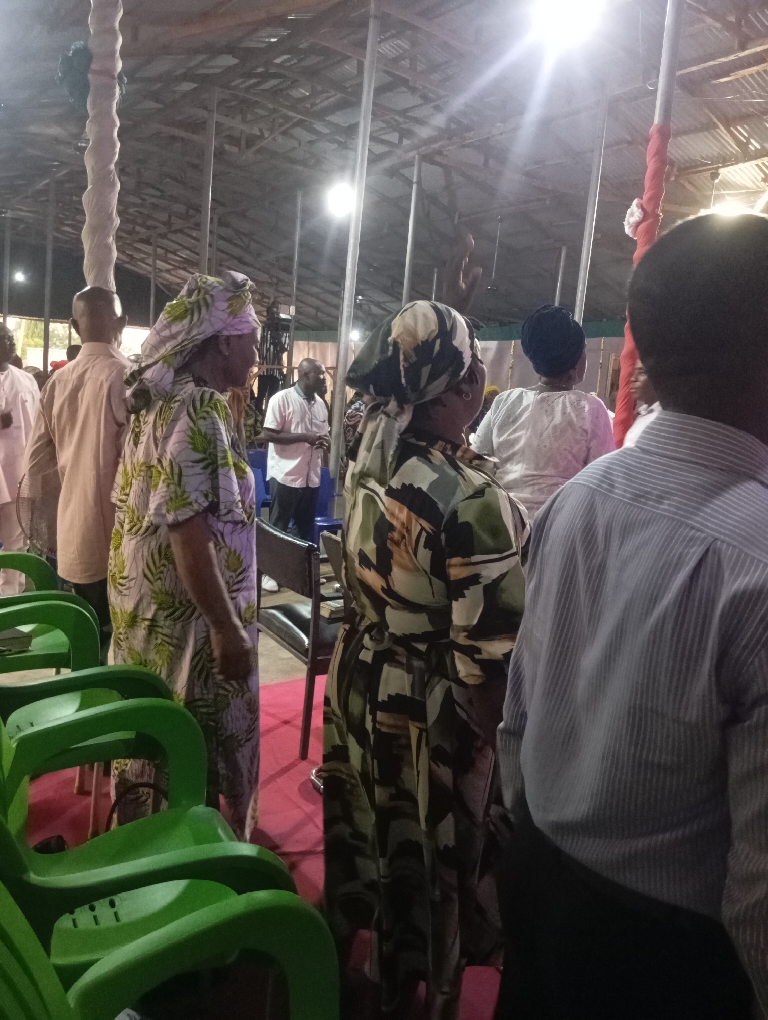 The photograph depicts a gathering of people indoors in what appears to be a church or meeting hall. The location is potentially in Markurdi, Nigeria. The structure has a corrugated metal roof supported by metal poles. The flooring is covered with red carpet. There are several green plastic chairs stacked in the foreground.
A group of adults are seen standing and looking towards a focal point, possibly a speaker or performer. Many are wearing patterned clothing and head coverings. Several men are dressed in shirts with collars.