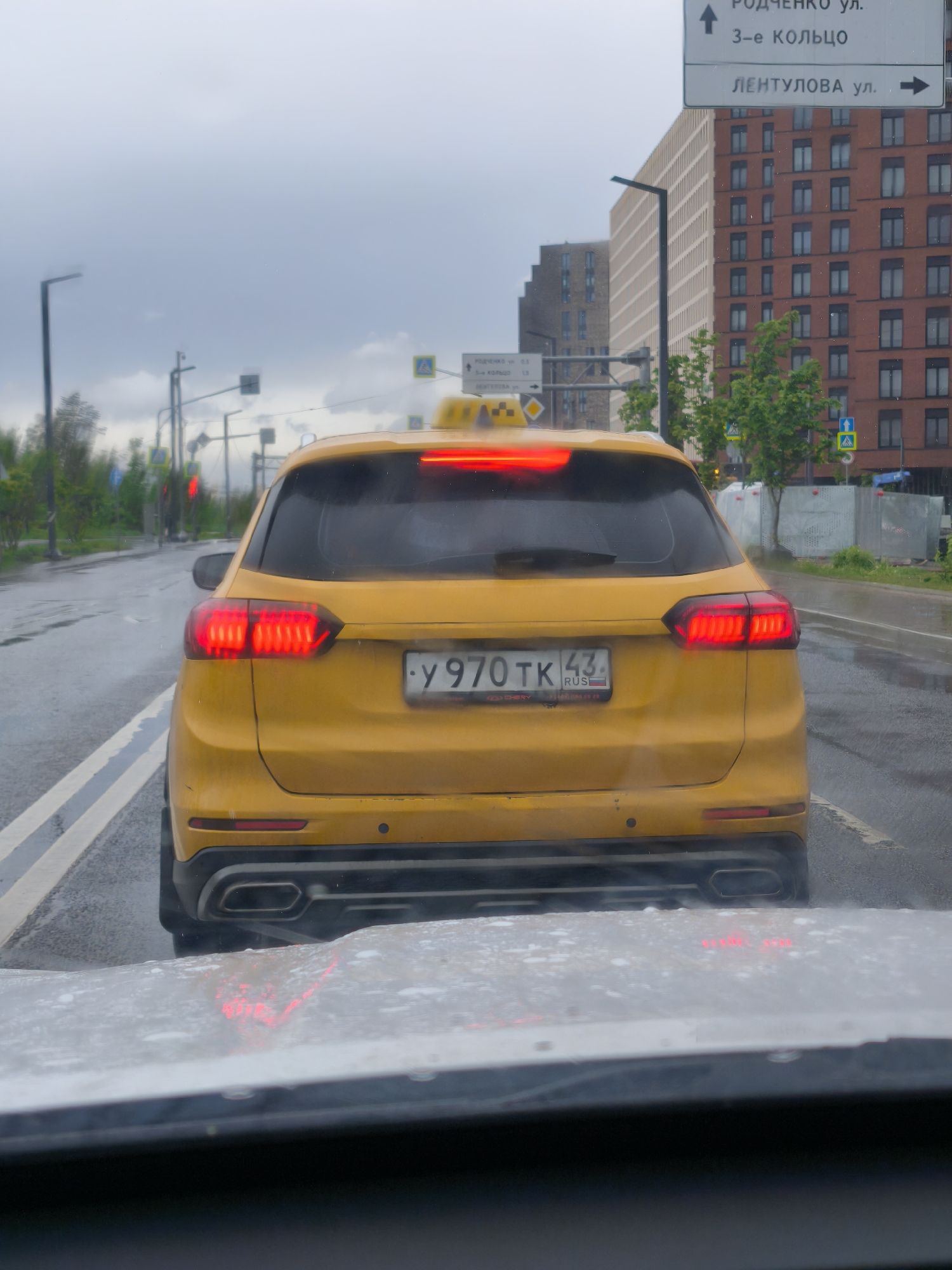 The image, taken from within a car, shows a yellow taxi in front, with its brake lights illuminated. The taxi's license plate is Y970 TK 43 RUS. Rain is visible on the windshield, and the overall weather appears overcast. In the background, there is a street sign indicating "РОДЧЕНКО ул." and "3-е КОЛЬЦО" above, with "ЛЕНТУЛОВА ул." pointing in the opposite direction. Several tall buildings are also visible, suggesting an urban setting. The sign and license plate both indicate that this is in Moscow, Russia.