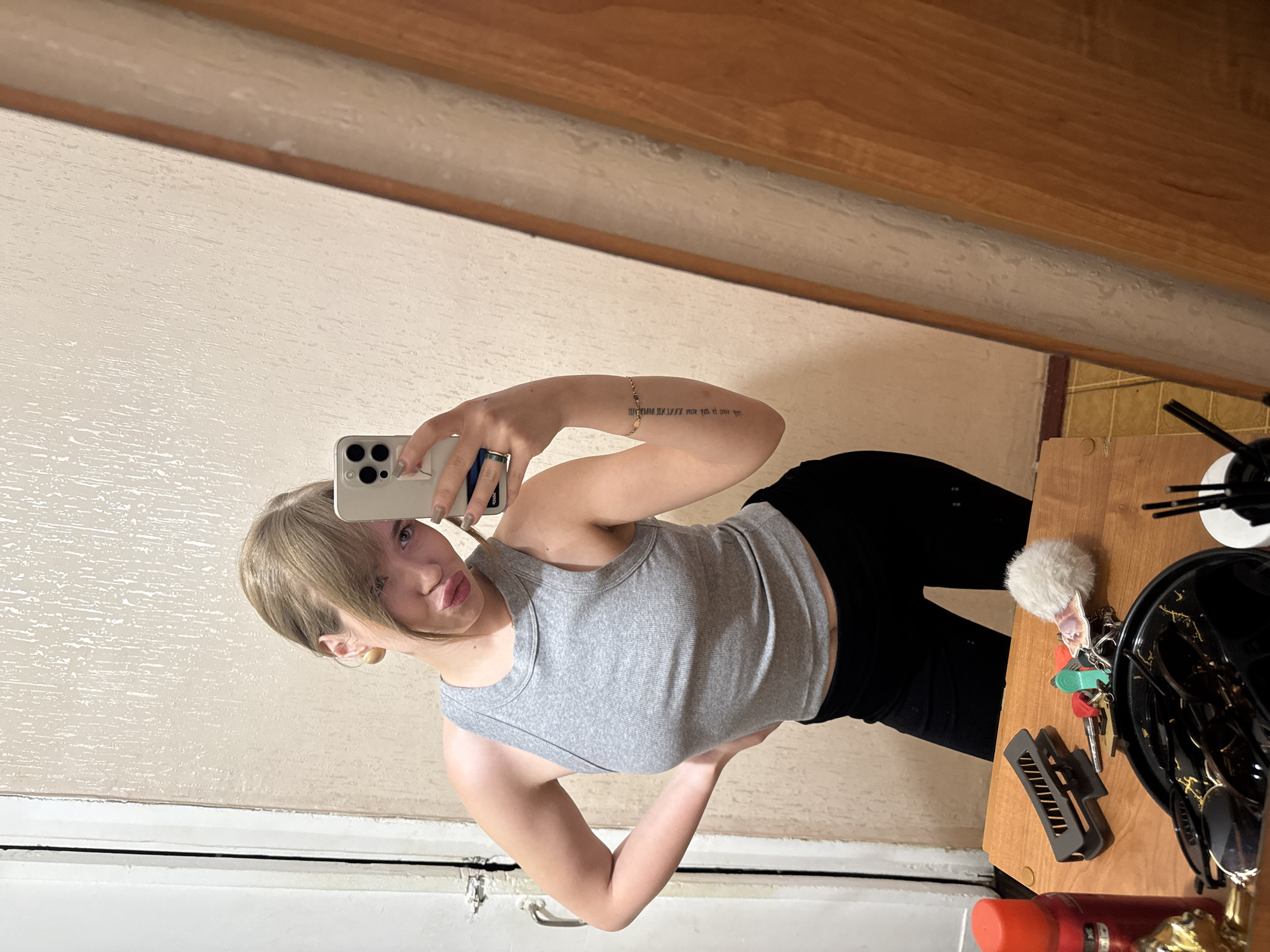 A woman is taking a selfie in front of a mirror. She is wearing a gray tank top and black pants. The background shows a room with light-colored walls, a wooden frame, and a wooden table cluttered with various items including keys, a hair clip, sunglasses, and a white fluffy keychain. It is not possible to determine the location as Chaikovsky, Russia, from the image.