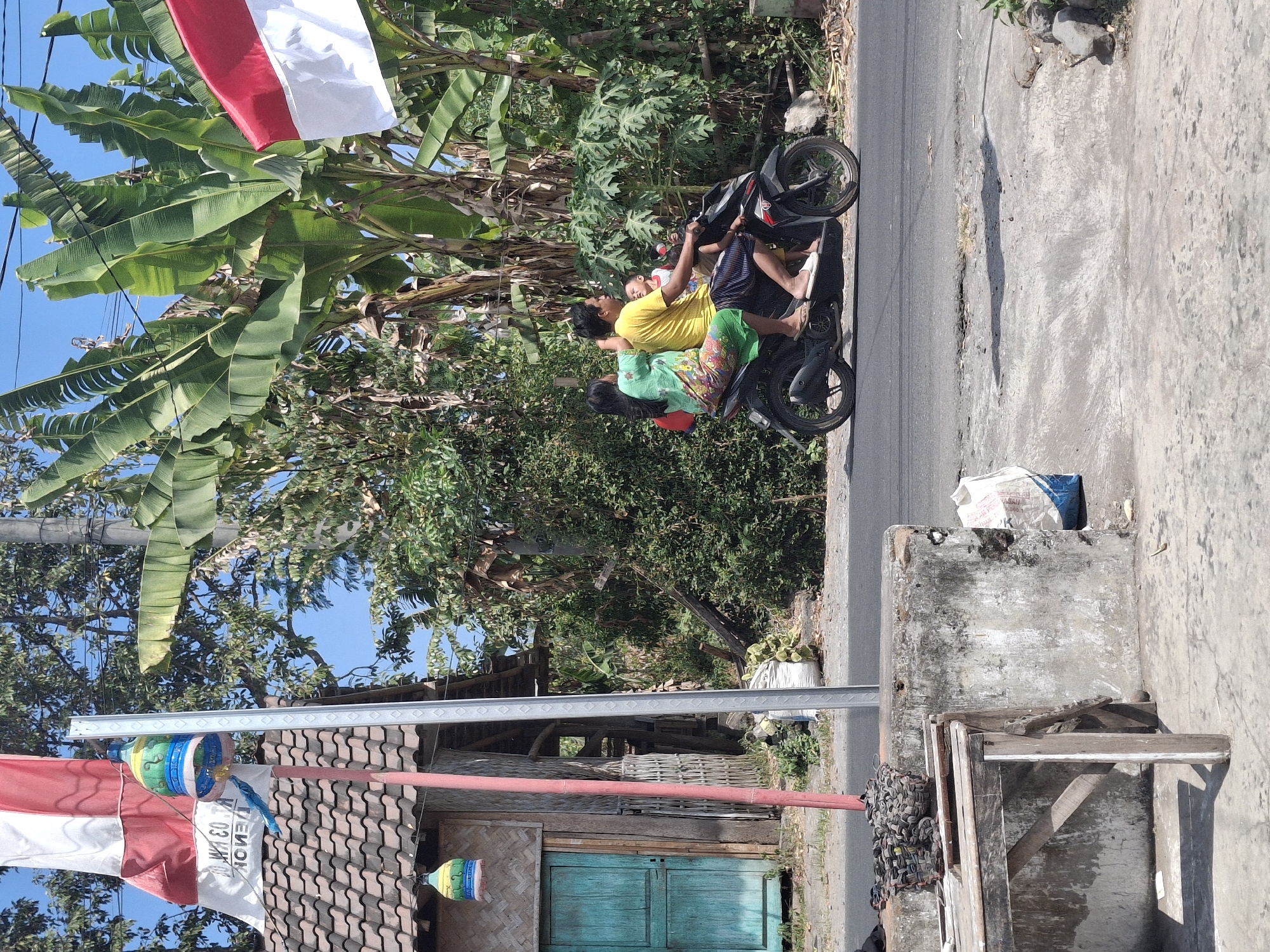 In Denok, Indonesia, on a sunny day, a family of four travels on a motorcycle along a paved road. The driver, a man wearing a yellow t-shirt and a sarong, is at the front. Behind him, a woman in a green and floral patterned dress holds a baby. Another child, in a red shirt, sits between the man and the woman. They are surrounded by lush greenery, including banana trees and other foliage. To the left of the road, a small, rustic building with a tiled roof and a turquoise door stands next to a flagpole flying a red and white Indonesian flag. Decorative birdcages hang from the pole and the building. In the foreground, a weathered wooden table rests on a concrete surface, with various objects, including what appears to be coiled rope, piled on top of it. A white bag is visible next to a concrete barrier in the background.