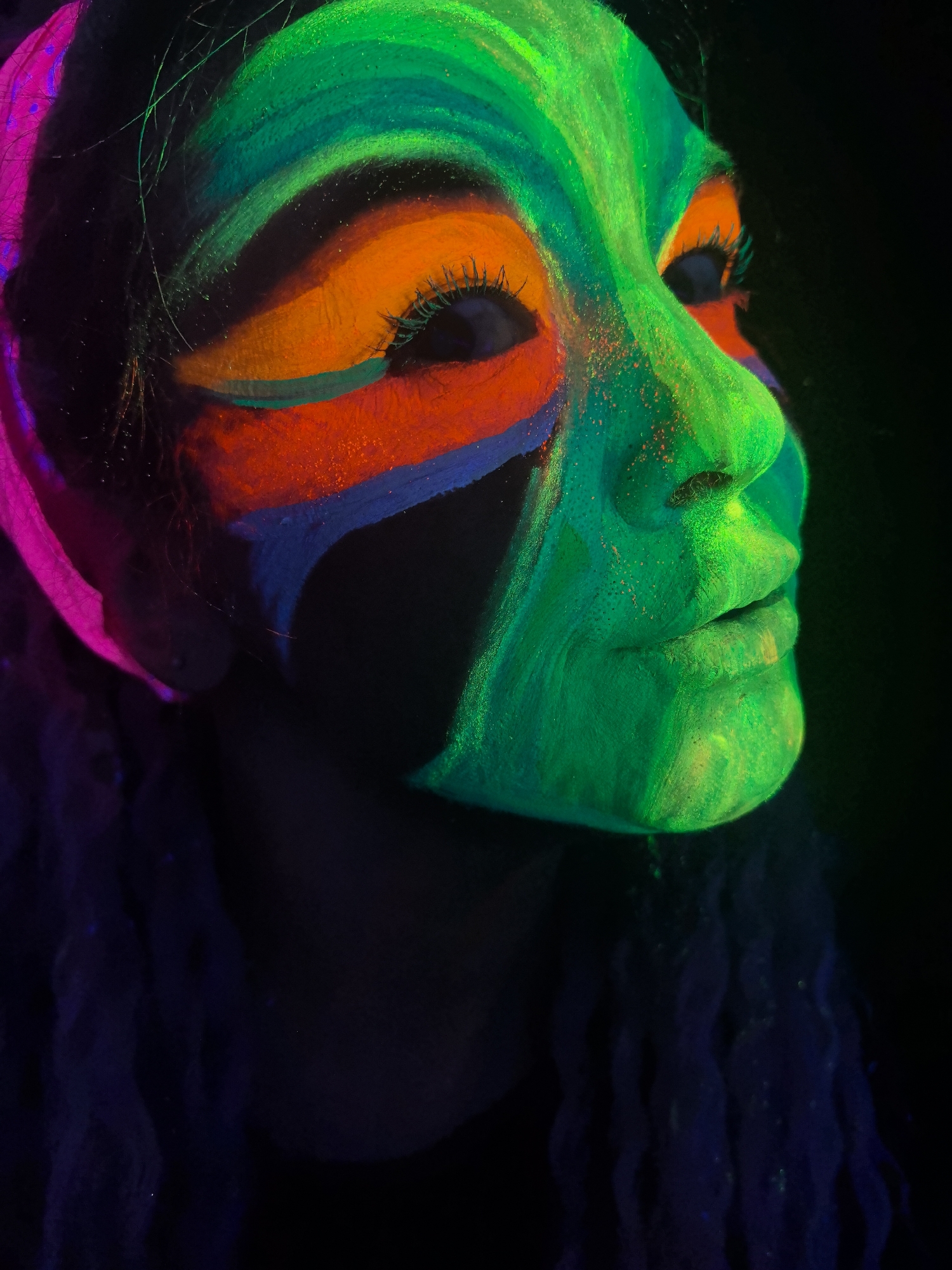 A close-up portrait shows a person with fluorescent face paint under ultraviolet lighting. The person's face is partially illuminated in shades of green, orange, red, and blue. Green covers the forehead, nose, and chin, while orange is painted around the eyes, followed by a stripe of red and blue. A portion of the head appears to be covered in bright pink. The person's eyes are visible, but the surrounding face is mostly black. The hair or clothing below the face is a dark, blurry color, and the background is black. There is no clear indication of location within the city of Noviy, Russia, or any other identifiable geographical or locational markers.