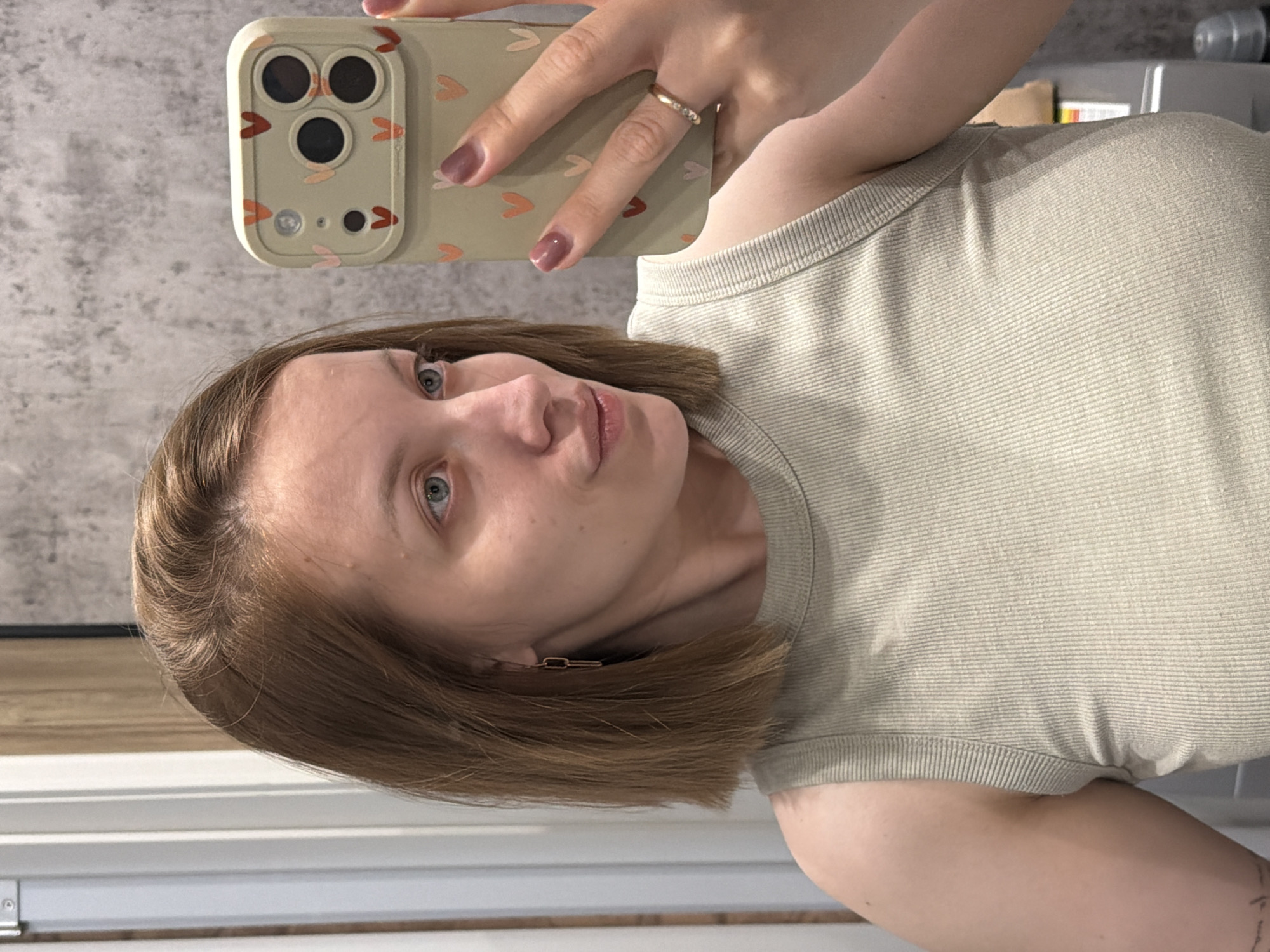 A woman with light brown hair and blue eyes is taking a selfie. She is wearing a light green ribbed tank top. Her phone case is light green with small orange hearts. She has a gold ring on her finger and a gold earring. She appears to be indoors, near a window. The background is a blurry grey wall. The location of Reshetnikovo, Russia is not discernible from the image.