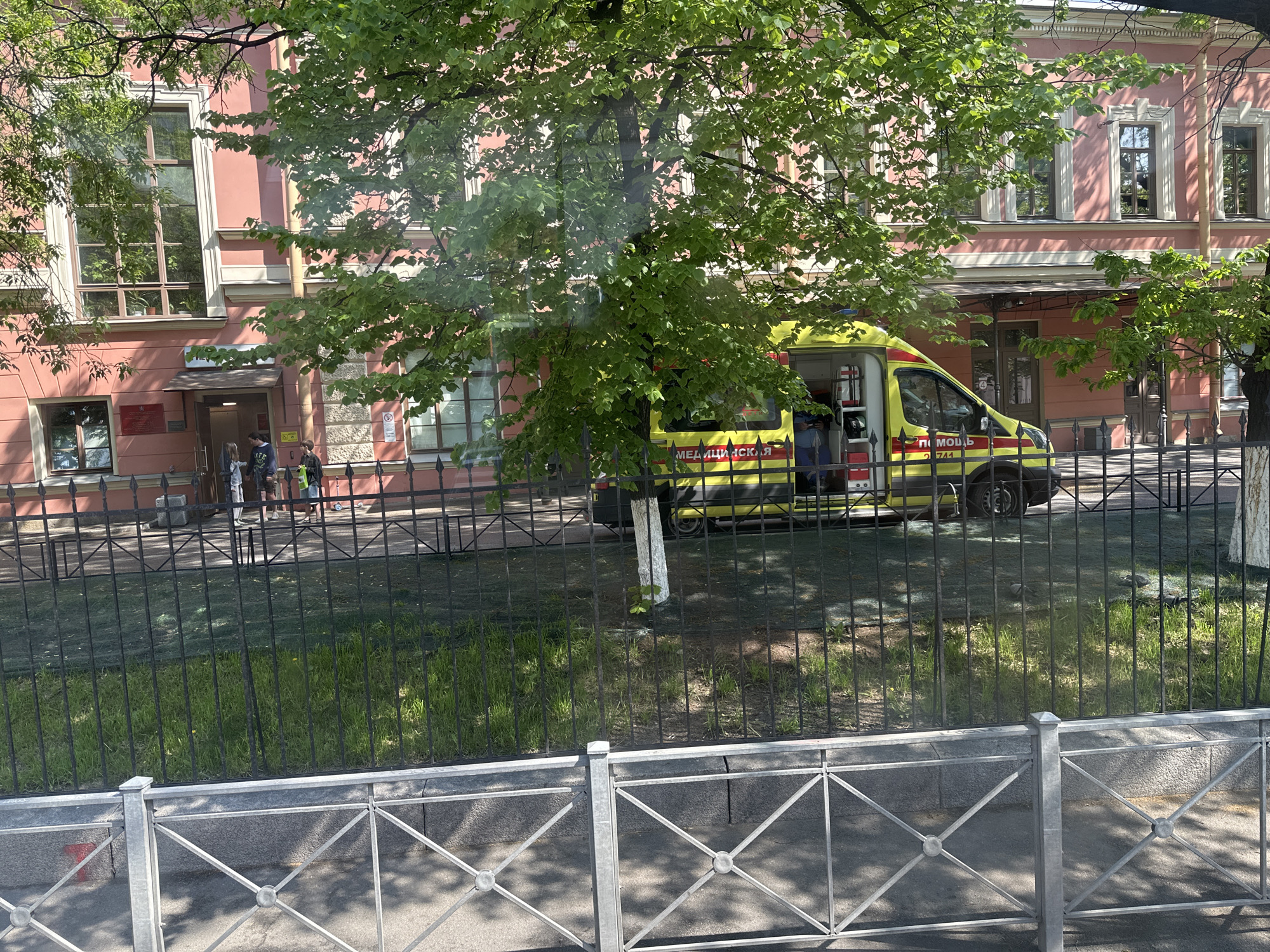 The image shows a pink building with multiple windows and entrances in Saint Petersburg, Russia. An ambulance van, painted in yellow and green, is parked in front of the building. A green tree partially obscures the view of the ambulance. A black metal fence separates the sidewalk from the building. People are seen entering the building. The image appears to be taken from behind a low, decorative metal fence.