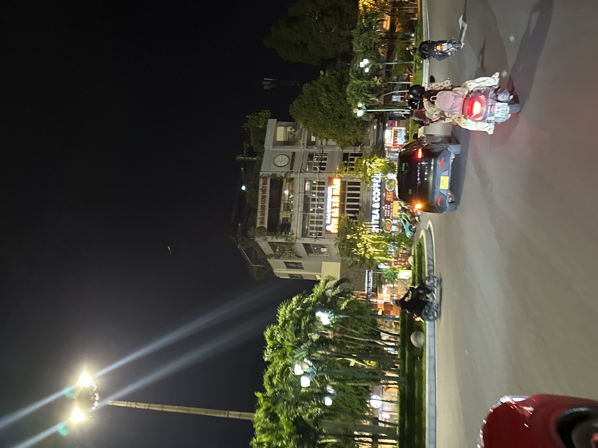 The image depicts a street scene at night in Nha Trang, Vietnam. A tall lamppost with multiple lights illuminates the left side of the frame, casting strong beams. Several palm trees are visible on the left, with streetlights interspersed among them. A multi-story building with signage is situated in the background. Signs on the building include "NHÀ HÀNG CƠM VIỆT NAM" and "TEA & COFFEE".
In the foreground, traffic is moving on a paved road. Several motorcycles are present. One motorcycle, ridden by a person wearing dark clothing and a helmet, is moving towards the left. Another motorcycle, carrying two individuals, one in a patterned dress with a backpack, is moving away from the viewer towards the right. A black SUV is stopped on the road, also facing towards the right. Another motorcycle is visible further down the road on the right, moving away. The scene appears to be a busy intersection or a section of a main road in the city. The overall lighting suggests it is nighttime, likely after sunset.