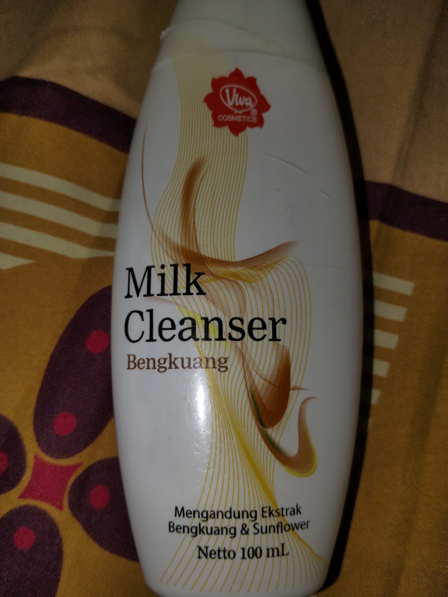 The image is a close-up, eye-level, indoor shot of a white plastic bottle of "Viva Milk Cleanser Bengkuang" standing upright. The bottle is adorned with a red Viva Cosmetics logo and stylized golden lines. The text on the bottle indicates it contains "Mengandung Ekstrak Bengkuang & Sunflower" and has a net content of "100 mL". The bottle rests on a textured surface with a pattern of alternating yellow and brown stripes and some darker circular motifs. The lighting appears to be artificial and somewhat dim, casting subtle shadows and highlighting the glossy surface of the bottle. There are no people or any elements suggesting a specific location in the city of Payagaboh, Indonesia. The scene is static and focused solely on the product.