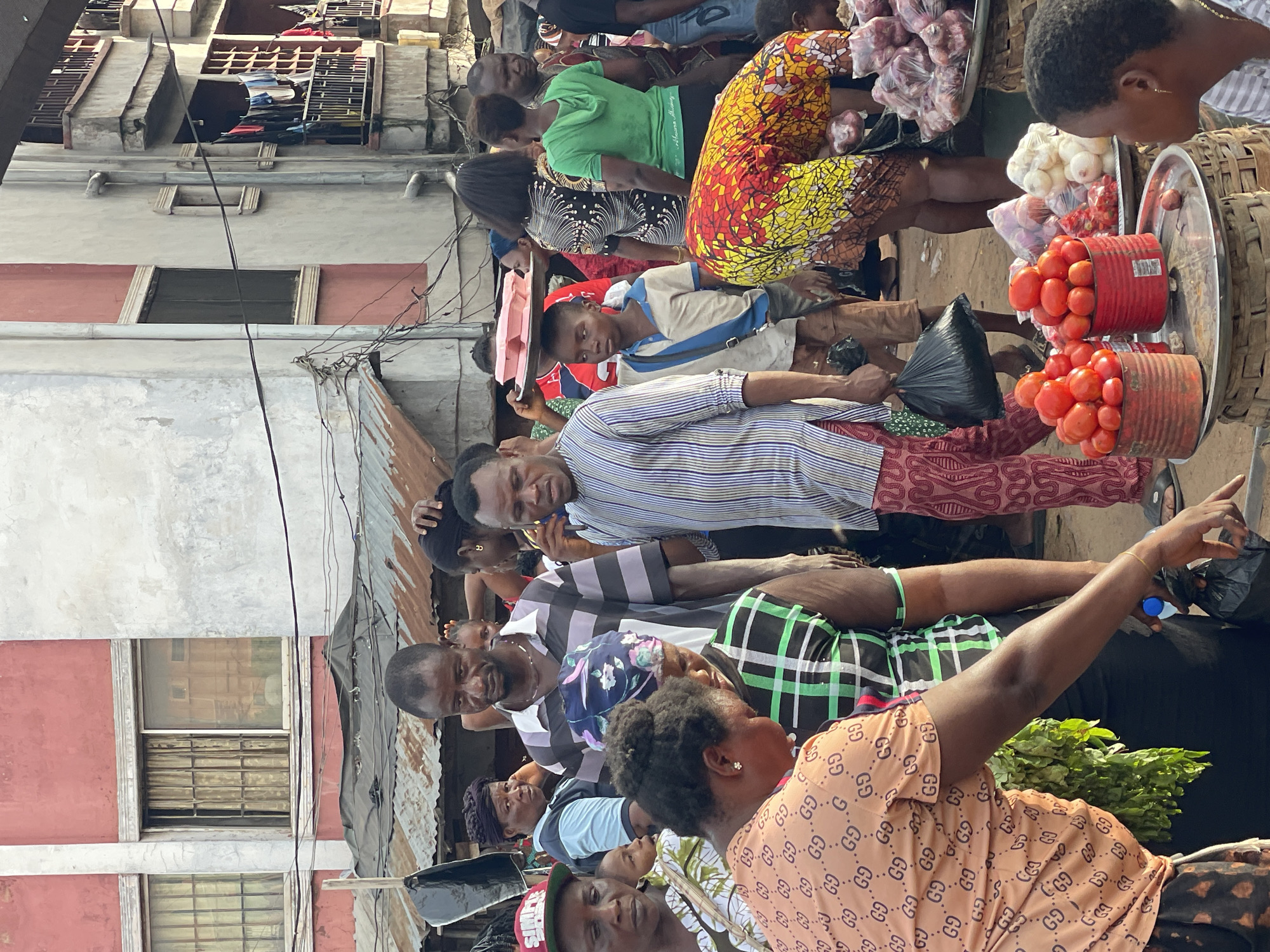 A medium, full-body shot shows a bustling market scene in Nkpor, Nigeria. People are densely packed, likely shopping for groceries, judging by the tomatoes, onions, and greens visible. One person is pointing with their right hand. Several are wearing patterned clothing; a woman is in an orange patterned shirt. Buildings are visible in the background, revealing an urban environment. The image is taken from a slightly elevated perspective.