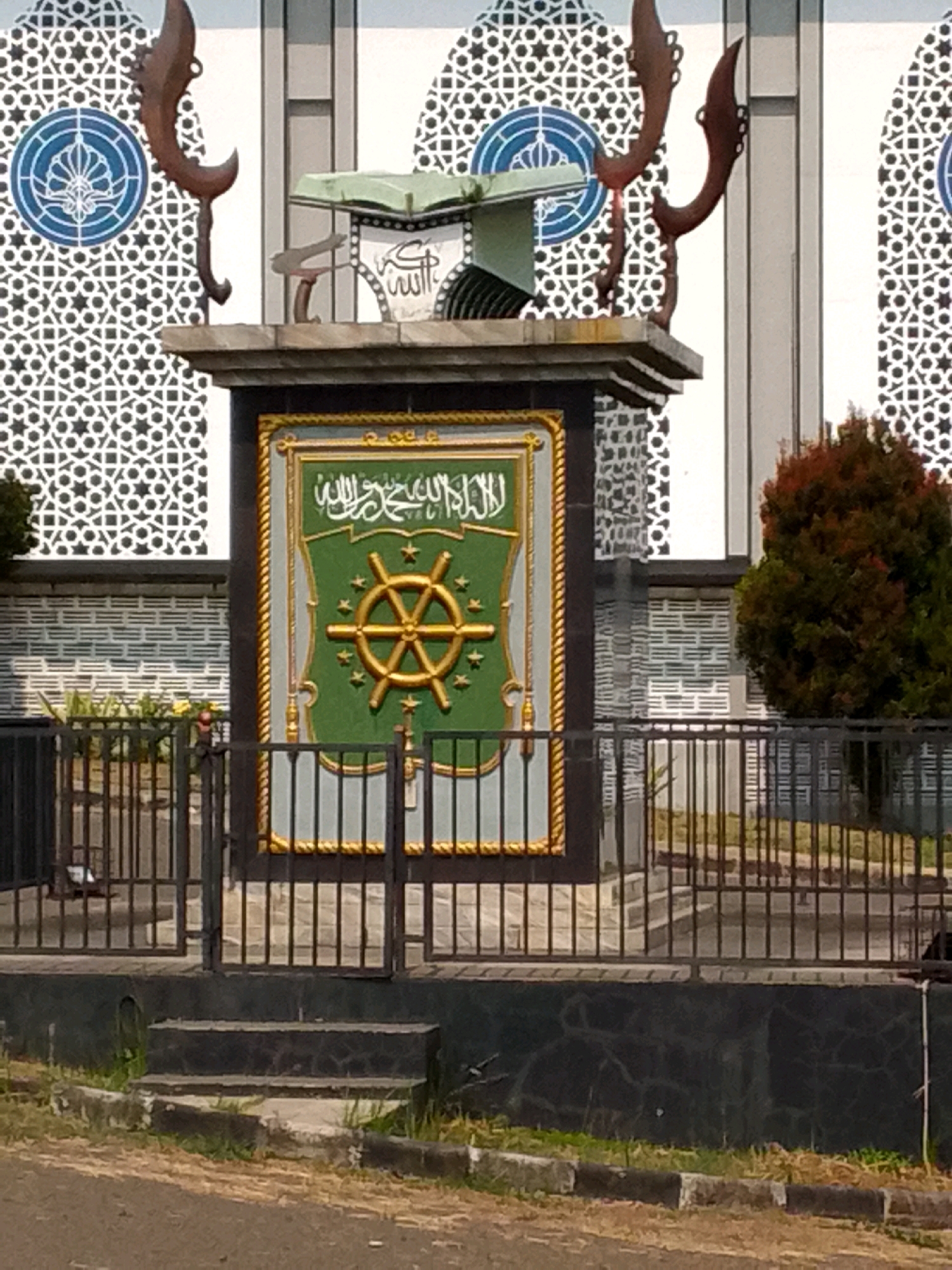 A large monument is pictured in Pasirhalang, Indonesia, with an open green book on top. The book is resting on a pedestal which has an emblem of a gold steering wheel with stars on a green background. The monument is framed by intricate Islamic patterns on the building behind it and a dark metal fence in front. A tree with red and green leaves stands to the right of the monument. The ground in front of the monument is paved with dirt and grass.