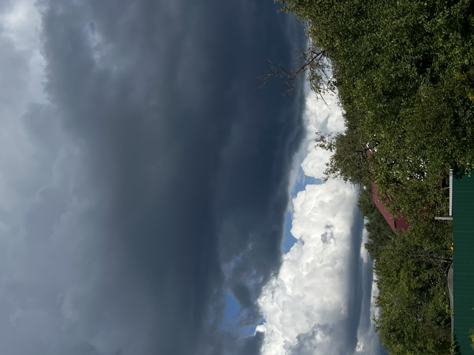 The image depicts an overcast sky with heavy, dark storm clouds dominating the upper portion. Below the storm clouds, lighter, fluffy white clouds are visible, interspersed with patches of blue sky. In the middle ground, lush green trees and foliage form a dense canopy. Partially visible through the trees is the red roof of a building, suggesting a residential or rural structure. A portion of a dark green fence runs along the bottom of the frame, separating the foreground from the scene beyond. The overall atmosphere suggests an impending storm or the aftermath of one. The location is identified as Strakhovo (Sitnikovskii S/S), Russia. There are no people or discernible actions or interactions visible in the image.