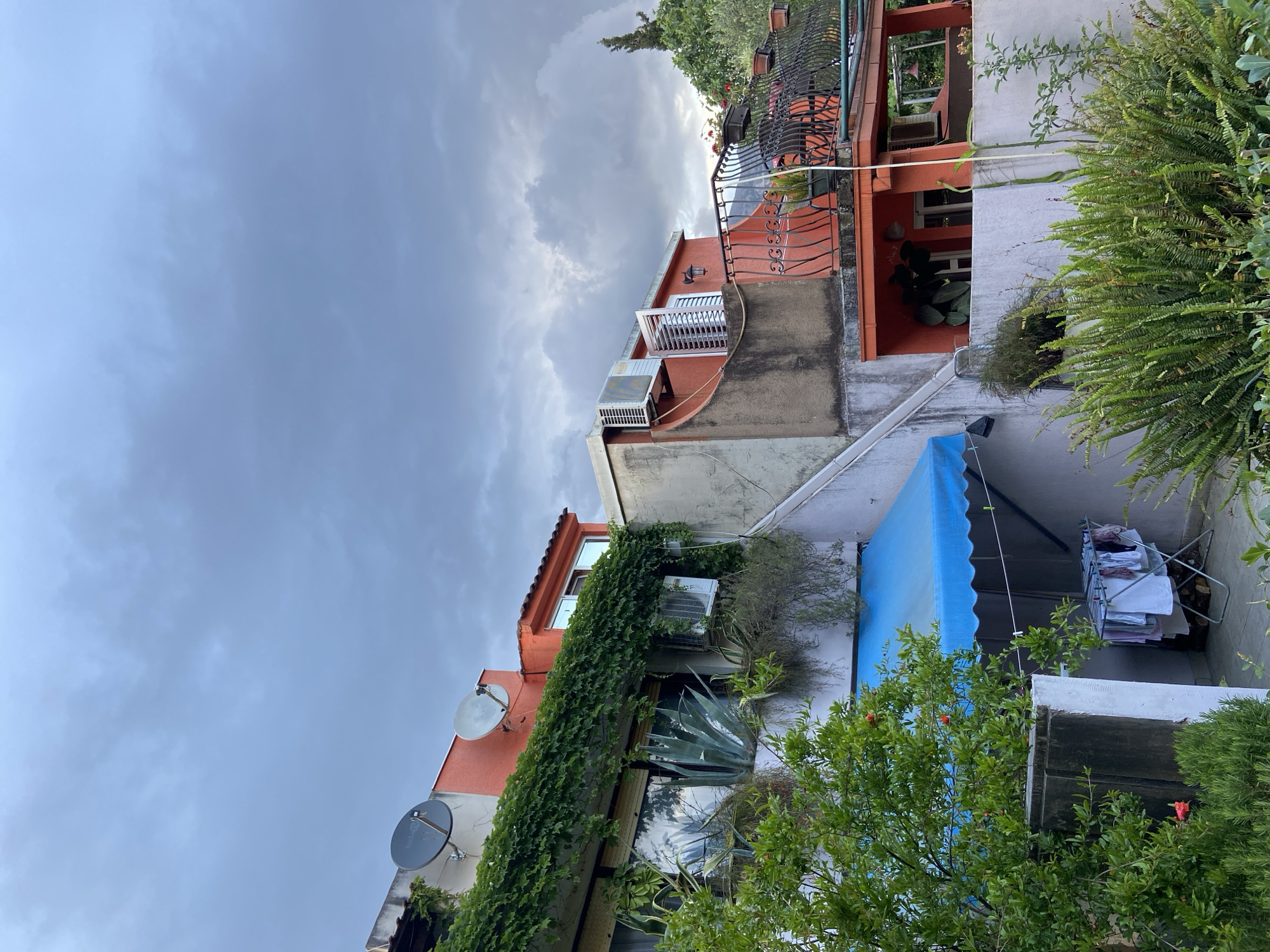 The image shows a street view in an urban location. The buildings are painted in shades of white, cream, and rust, with some areas covered in ivy. There are satellite dishes mounted on some of the buildings, and a blue awning extends over a ground-level patio. The sky is overcast with dark clouds. The overall impression is of an older, densely built area with a mix of residential and possibly commercial properties. The location in the city of Unknown, Unknown is provided as reference, but there are no specific indicators in the image to verify this location.