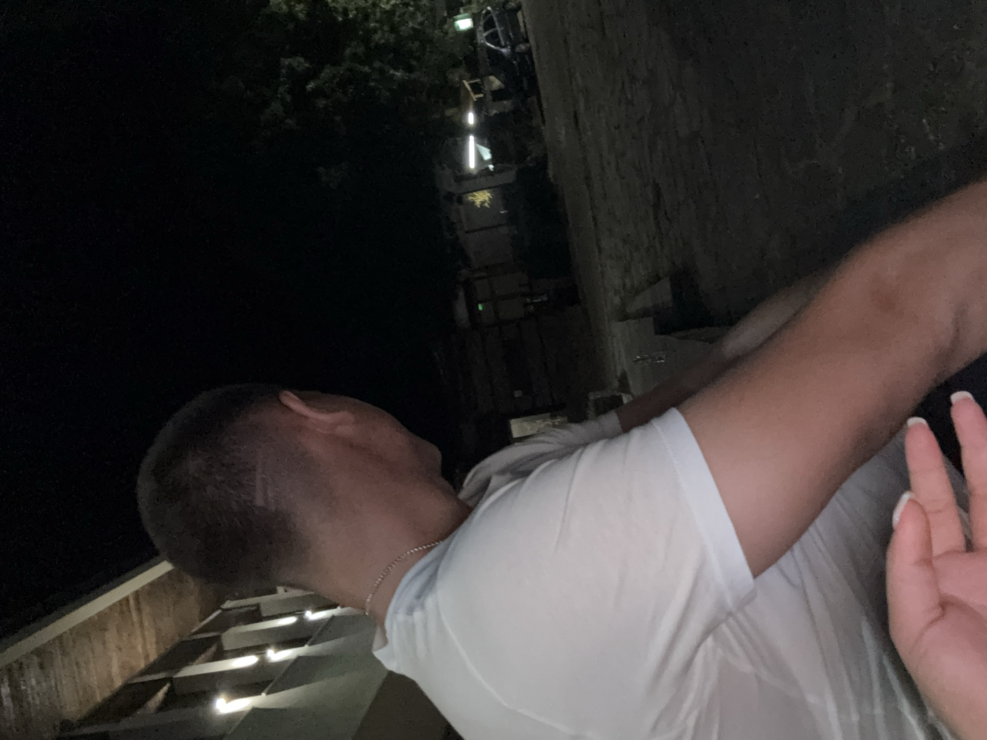A man wearing a white t-shirt and a necklace is shown from the shoulders up. His hair is short and dark, with a visible line design at the back of his head. He is facing away from the camera, looking to the right. In the background, there is a dark outdoor environment with some trees and buildings. The scene appears to be illuminated by artificial lights, suggesting it is nighttime. A car is parked in the distance. The image is taken in the city of Sopot, Bulgaria.