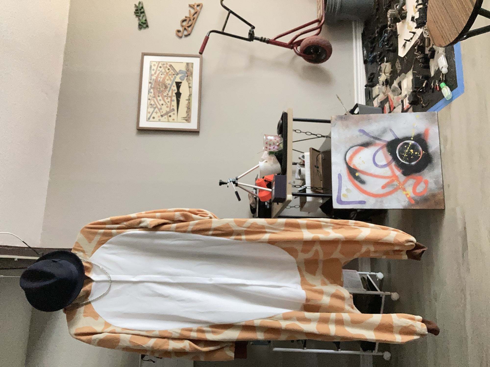 The image depicts an indoor scene, possibly a residential room or a studio space. A giraffe-patterned onesie with a white chest panel is hanging from an unseen support, with a dark fedora hat placed on top of the onesie's hood. A silver chain necklace is draped around the neck area of the onesie.
To the right, a small red scooter with a black handlebar and red grips is leaning against the wall. A framed picture hangs on the wall above the scooter, depicting a colorful, abstract design with geometric shapes and figures. Two decorative wooden pieces with letters are also mounted on the wall, one reading "SEKA".
In the mid-ground, a black metal table with a wooden top holds a black tripod, a white cup, and a small glass with a pink pattern. To the right of the table, a gray bucket is visible.
In the foreground, a square canvas with spray-painted abstract art is placed on the floor. The art features orange, purple, and black spray paint. The floor is cluttered with various items, including what appear to be electronic components, wires, and other debris, some of which are arranged on a black mat outlined in blue tape. A round wooden table is partially visible in the lower right corner. The room has light-colored walls and a concrete or linoleum floor. The overall impression is one of an artistic or somewhat disorganized personal space.
This image was captured in Round Rock, United States of America.