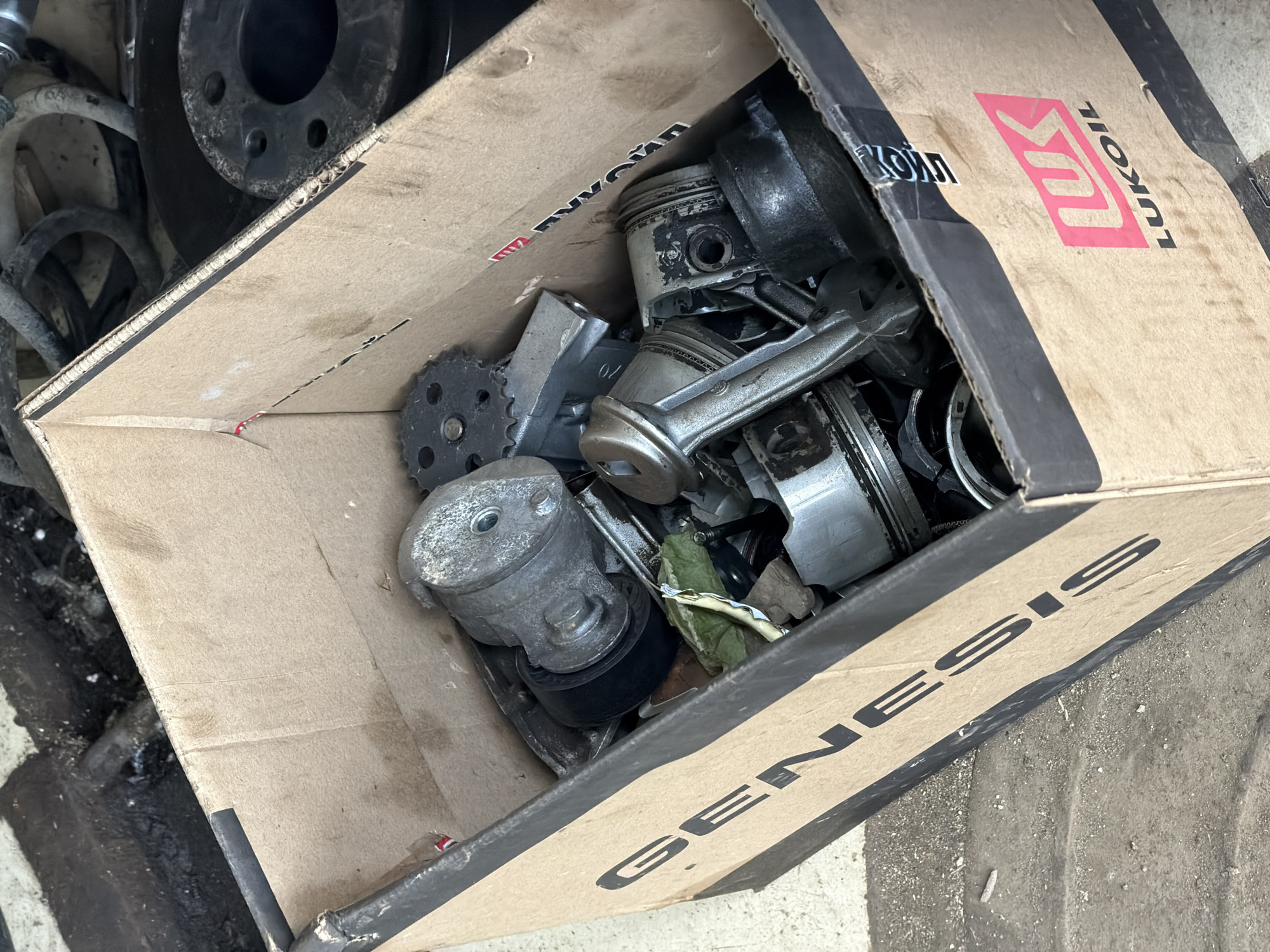 A cardboard box, labeled "GENESIS" and featuring a "LUKOIL" logo, is filled with various engine parts, including pistons, a crankshaft, a gear, and a pulley system. The box rests on a concrete or dirt floor, with some debris scattered around it. The scene appears to be indoors, possibly in a garage or workshop, given the presence of oily residue on the box and the general appearance of the environment. There are no people visible in the image. The image is a close-up shot, focusing on the contents of the box and its branding.