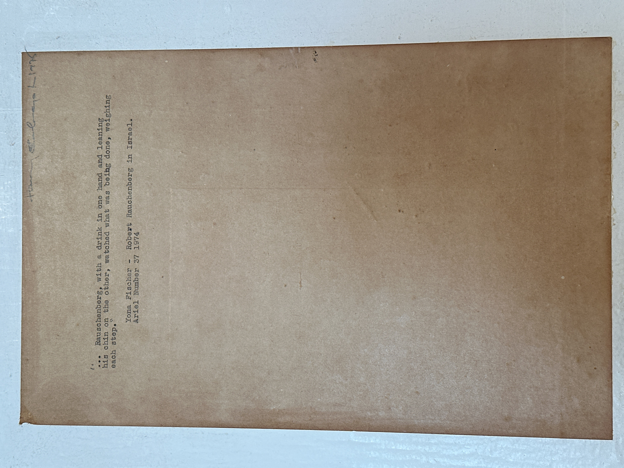 The image shows a typed description of Robert Rauschenberg in Tel Aviv, Israel. The description reads: "...Rauschenberg, with a drink in one hand and leaning his chin on the other, watched what was being done, weighing each step." It is credited to Yona Fischer in Ariel Number 37, 1974. Handwritten in the top left corner is the year "1975". The text is typed on a sheet of brown paper.