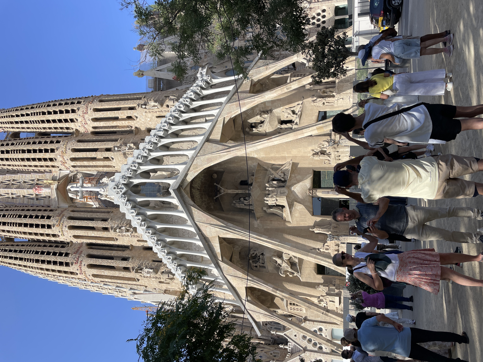 The image shows a sunny daytime exterior view of the Sagrada Familia basilica in Barcelona, Spain. The iconic, unfinished basilica dominates the background with its intricately carved stone facade and soaring spires. In the foreground, a group of tourists are gathered, some are looking at the basilica, some are taking pictures, and others are interacting with each other. The ground appears to be a dirt or gravel path. Trees with green foliage are visible on the left and right sides of the image, providing some shade. The sky is a clear, vibrant blue. The overall impression is a tourist gathering at a famous landmark on a bright day.