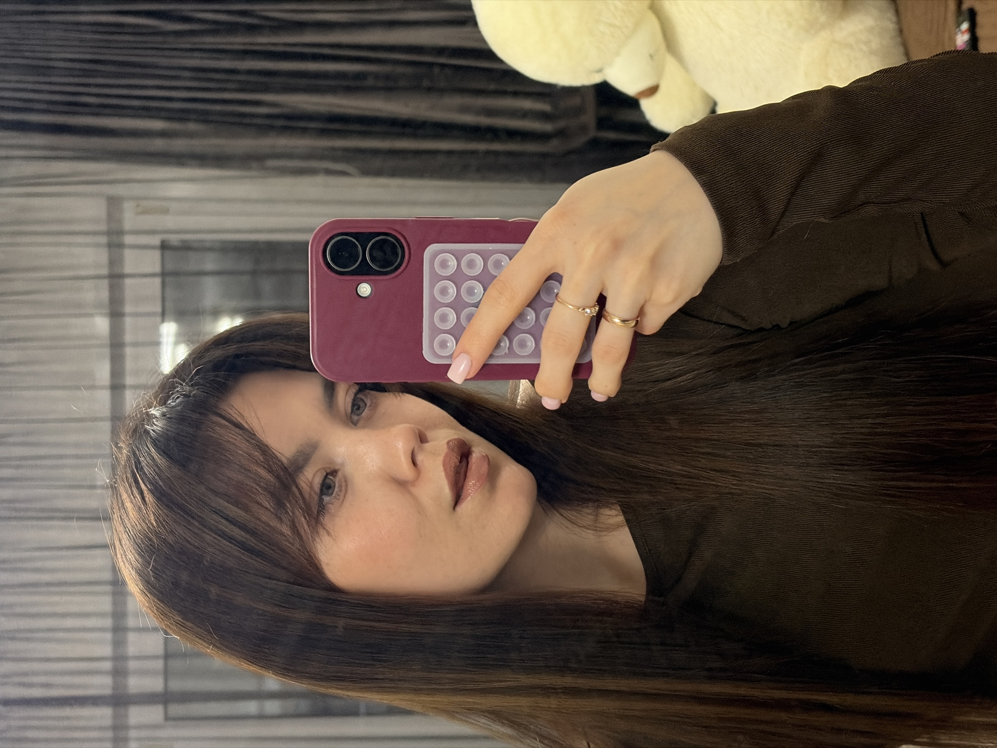 A selfie is taken indoors by a woman with long brown hair and light skin. She holds a maroon cell phone case with a grid of small, clear suction cups and two camera lenses. Two gold rings are visible on her fingers. The background includes a window with horizontal blinds or a similar pattern, and part of a soft, cream-colored teddy bear. The location in the city of Votkinsk, Russia is unknown.