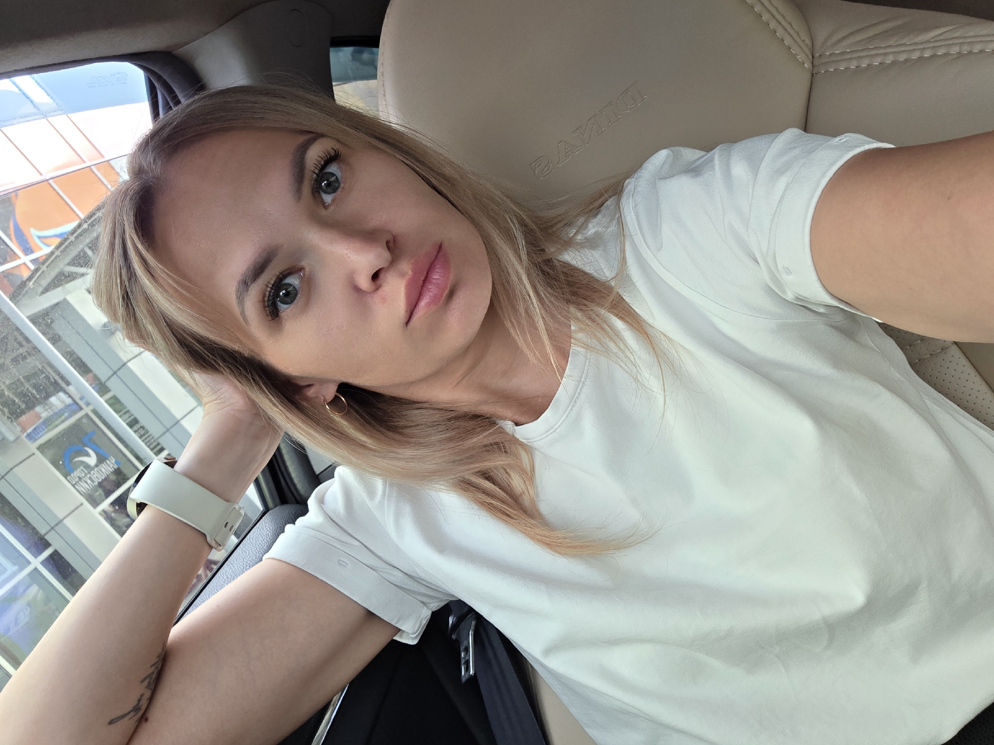 A woman with blonde hair and blue eyes takes a selfie while sitting in the passenger seat of a car. She is wearing a white t-shirt and a white smartwatch on her left wrist. Her right arm is resting on her head. The car's interior is visible, including a beige leather seat and a black seatbelt. Outside the car, a building with reflective windows can be seen. The scene appears to be in the city of Chaikovsky, Russia, as indicated by text on the building visible in the background.