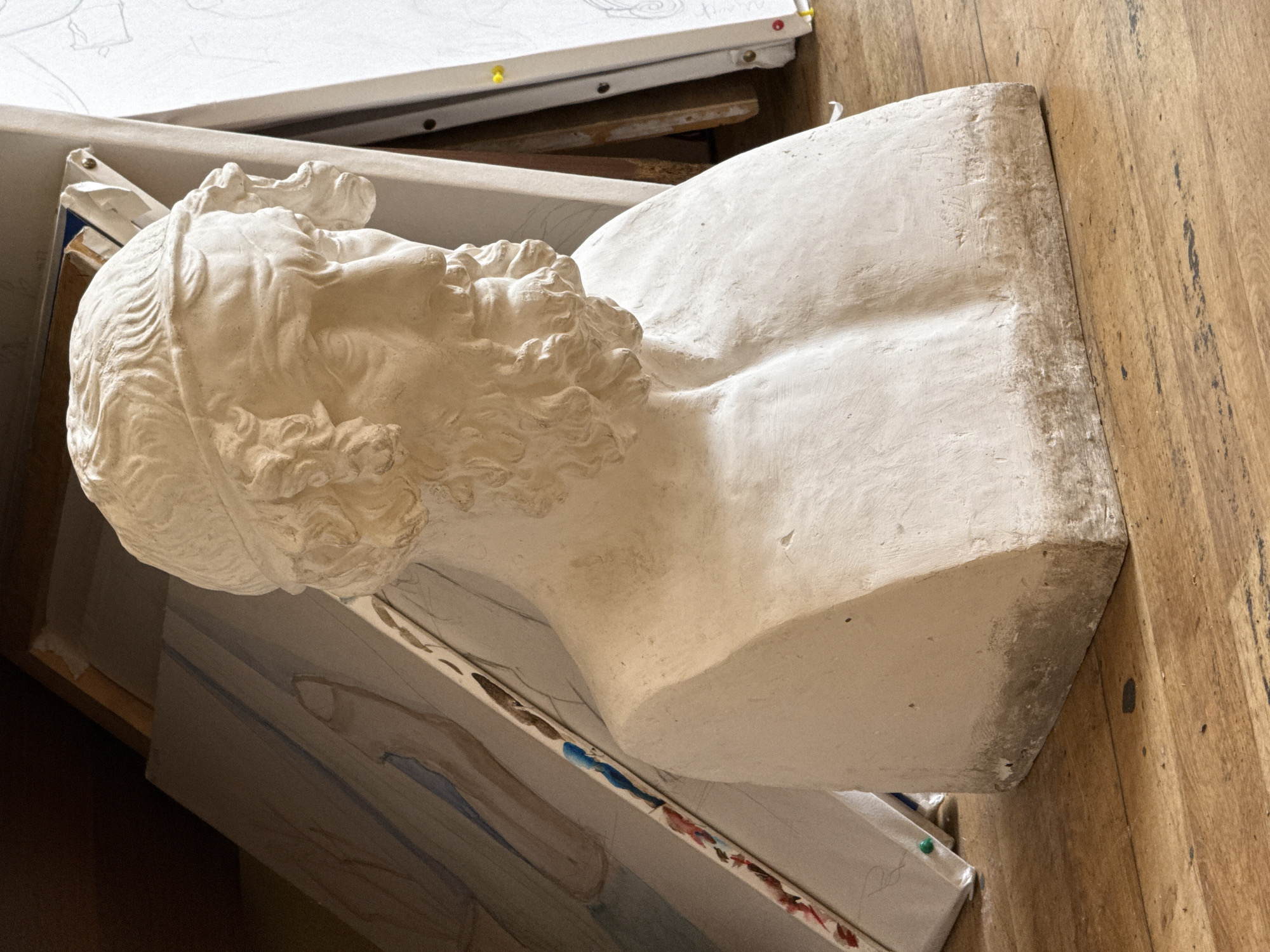 The image shows a plaster bust of a bearded male figure, possibly a classical or neoclassical sculpture, resting on a stack of canvases in a studio setting. The bust has detailed curly hair and beard. The wooden floorboards are visible in the background. The exact location in Yerevan, Armenia, is not discernible from the image. There is no mention of any people in the image.