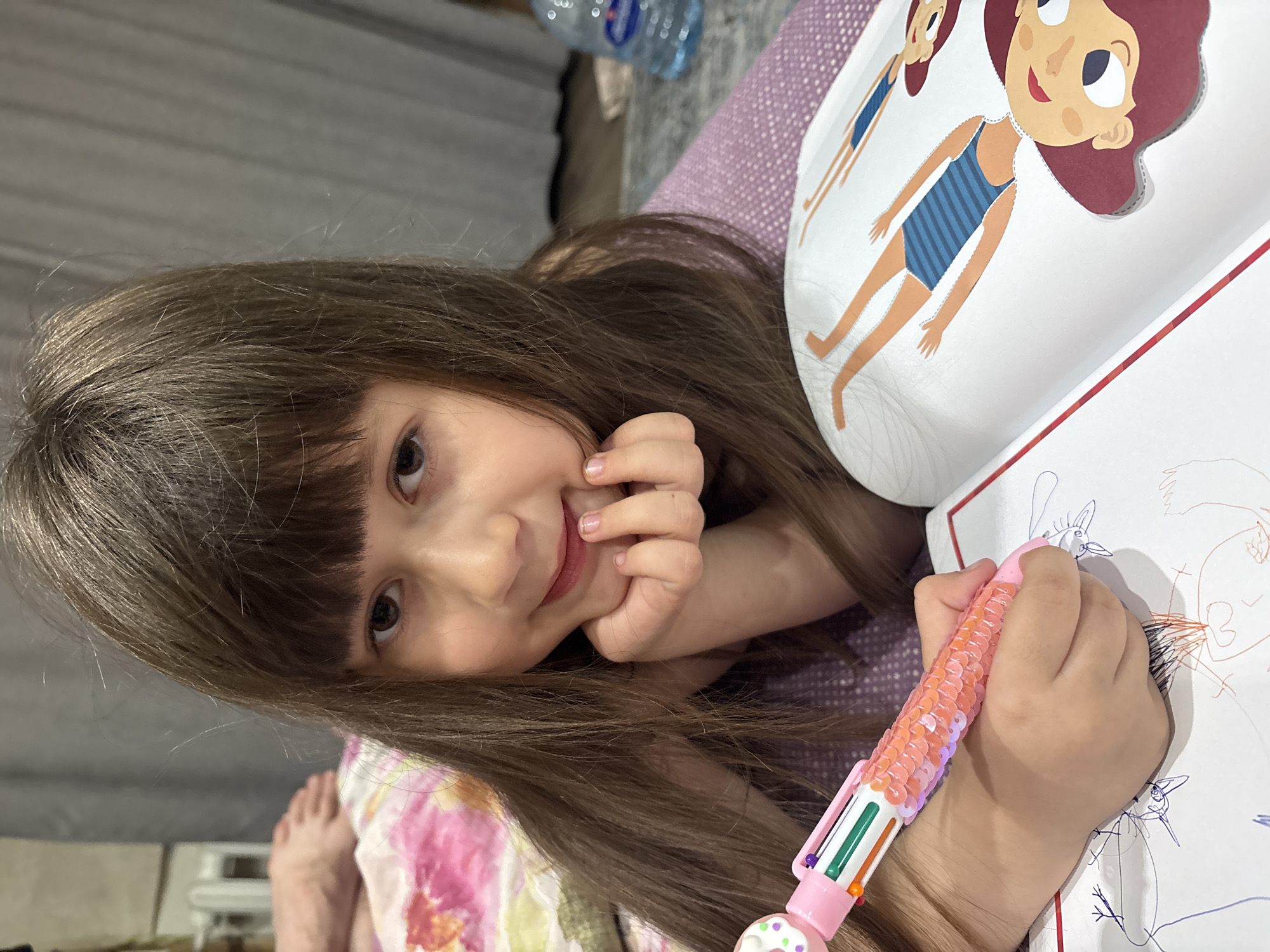 A young girl with brown hair and bangs is lying on a surface with a floral print, possibly a bed or couch. She is holding her hand to her cheek and looking at the camera with a slight smile. In her other hand, she holds a multi-colored pen and is drawing on a piece of white paper with scribbles. A book is open beside her with a cartoon image of a girl in a blue and white striped swimsuit. A bottle of water is visible in the background. The location is not discernible from the image.