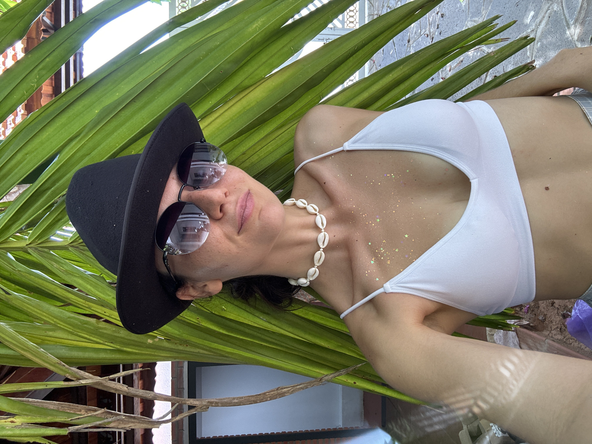 A woman wearing a black fedora, round sunglasses, and a white bikini top stands behind a palm frond in Mũi Né, Vietnam. She is adorned with a shell necklace and glitter on her chest. The background shows a building with a tiled roof and a decorative fence. The lighting suggests it is daytime.