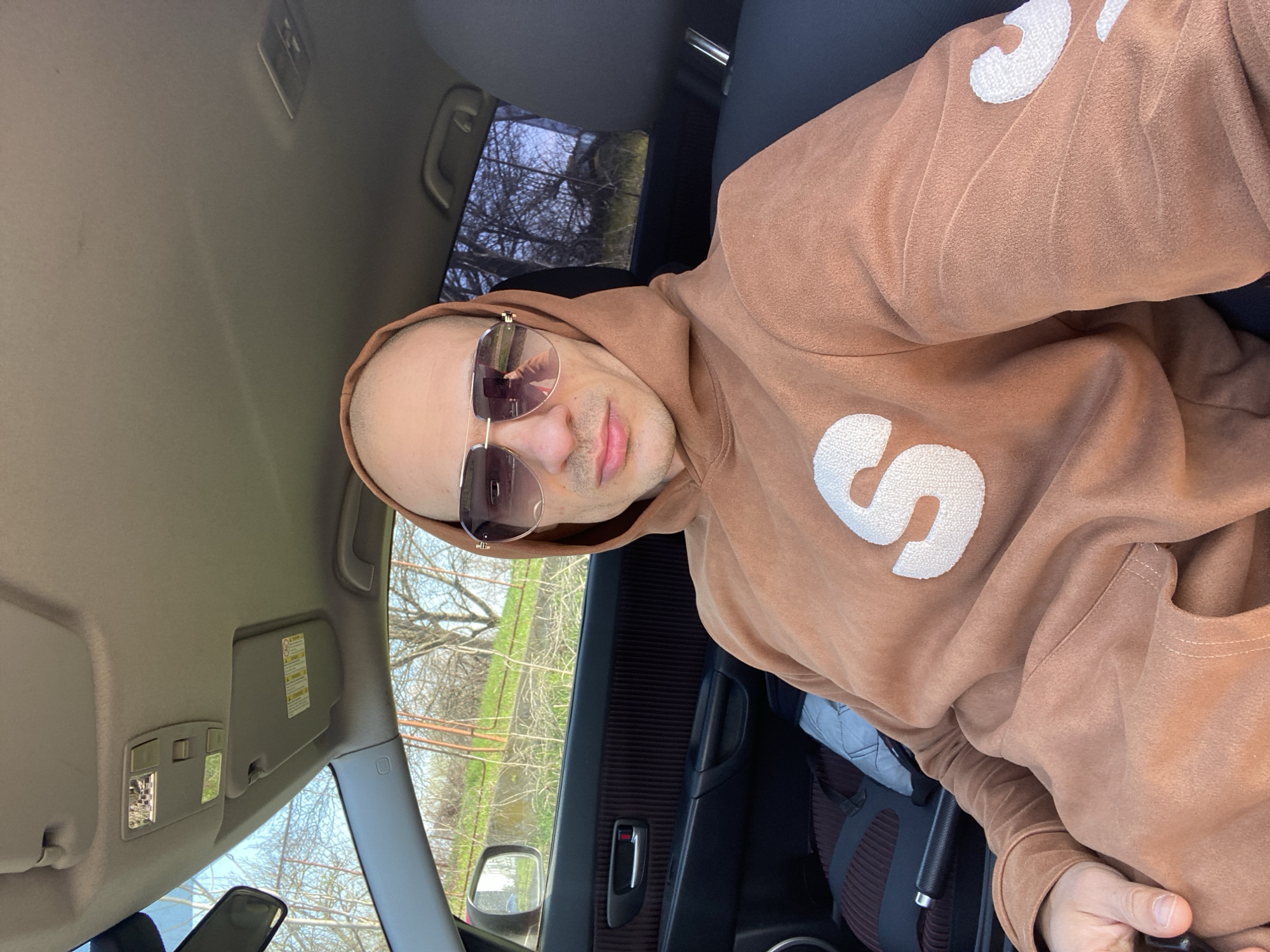 A man is seated in the driver's seat of a car. He is wearing a light brown hoodie with a large white "S" on the front and sunglasses. The car's interior is visible, including the dashboard, door panel, and seats. Outside the window, trees and greenery are visible, and based on the location request, the scene may be within the city of Pleven, Bulgaria.