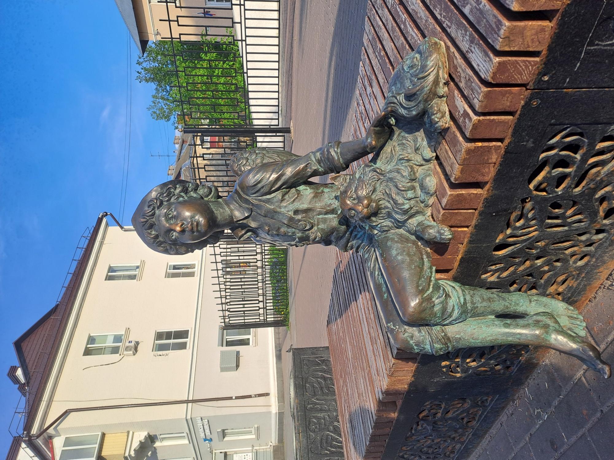 A bronze statue of a young boy sitting on a wooden bench with a dog is the central element of this photograph taken in Elektrostal, Russia. The statue is positioned outdoors, with a multi-story building and a black wrought-iron fence visible in the background. The day appears sunny, as indicated by the bright blue sky. The bench has a dark, decorative metal base.
