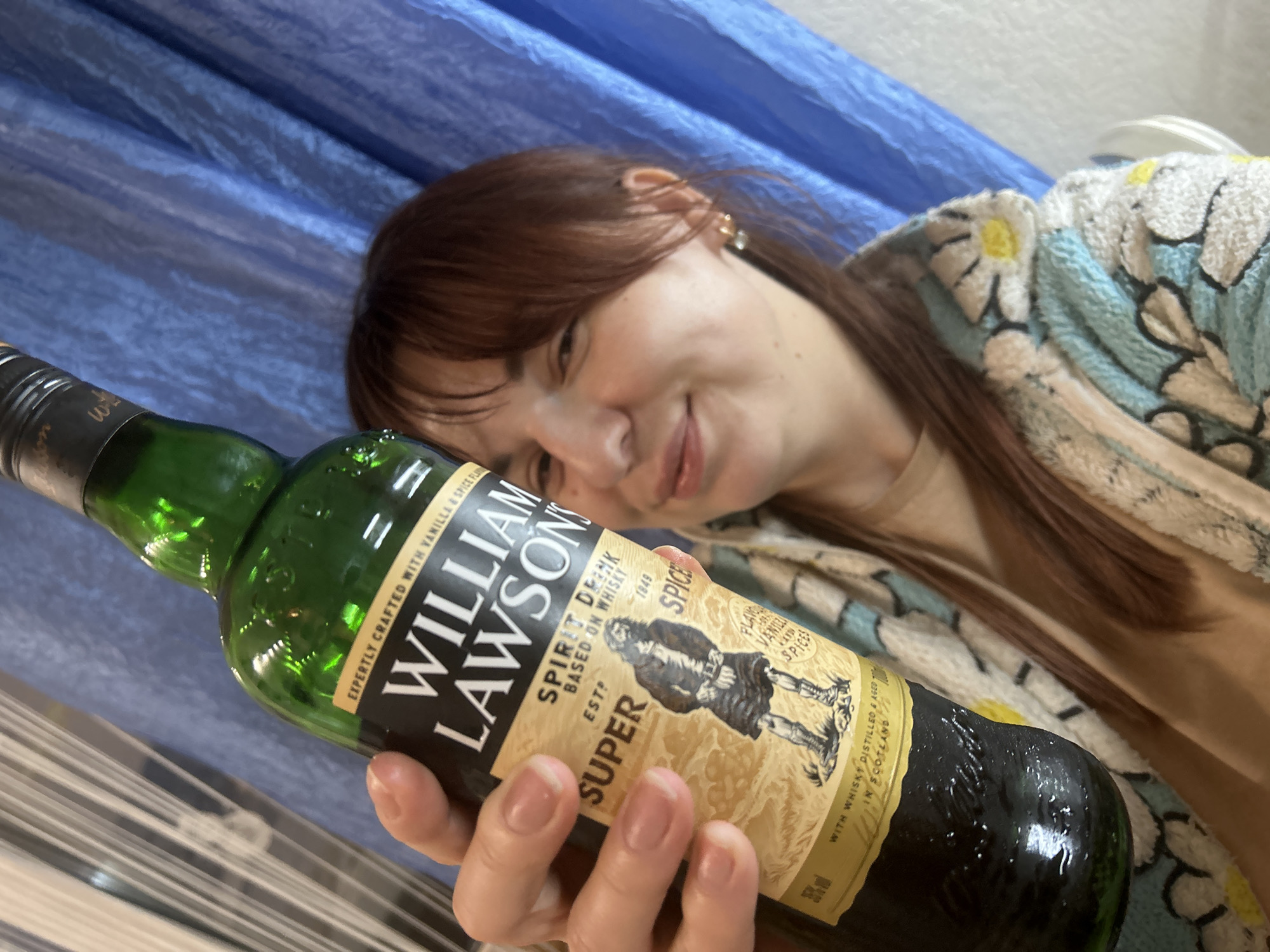 A woman with reddish-brown hair is holding a green bottle of "William Lawson's" Spirit Drink based on whisky, Super Spice edition, in front of her. The bottle's label is partially visible. The woman is smiling and wearing a light-colored robe with a floral pattern. A blue curtain is visible in the background. The image provides no information about the location in the city of Фоки, Russia.