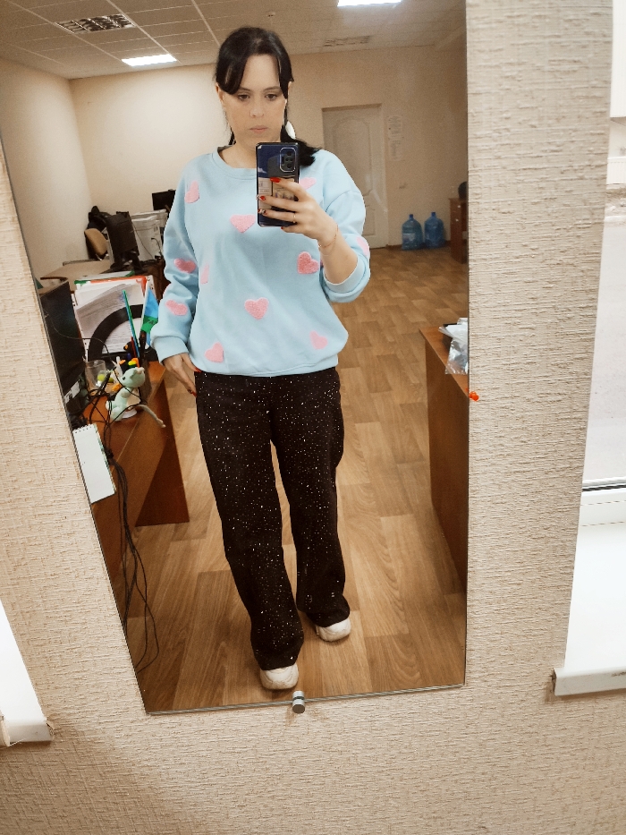 An adult female is depicted in a full-body mirror selfie, standing indoors. She has dark hair, partially pulled back, and wears a light blue long-sleeved sweatshirt adorned with multiple pink heart-shaped appliques. She is also wearing dark trousers with a speckled pattern and white sneakers. Her right hand holds a dark-colored smartphone, positioned to capture her reflection, with a thin bracelet visible on her right wrist.
The setting is an interior room, likely an office or common area. The walls immediately surrounding the mirror are covered with textured, light beige wallpaper. The room visible in the reflection features plain, light-colored walls and a light brown laminate or wood-patterned floor. The white ceiling contains multiple recessed rectangular fluorescent light fixtures.
In the background reflection, to the left, a wooden desk is visible with several items including what appear to be computer monitors, tangled cables, a white notebook or binder, and a green plush toy. To the right, further back, another wooden desk is present, and on the floor beneath it, two large blue plastic water bottles are placed. A light-colored door is visible slightly ajar in the far background.
The exact location within the city of Donetsk, Ukraine, cannot be determined from the visual information provided in the image.