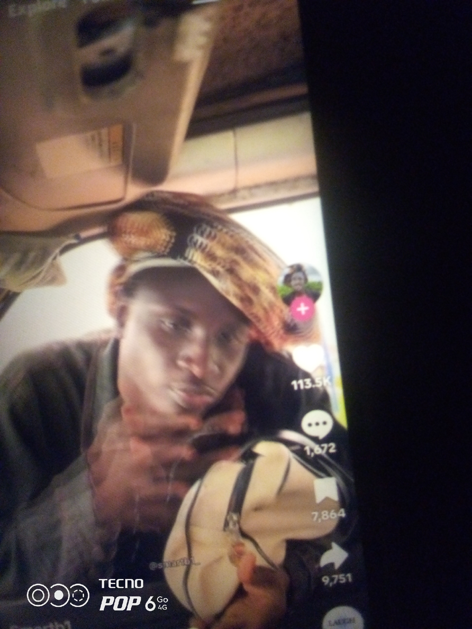 The image is a low-resolution photograph, likely taken from a mobile device, displaying a person from the chest up. The person is wearing a hat with a patterned brim and appears to be inside a vehicle, possibly a truck, given the visible roof structure and a label on the visor. The background is blurry, but shows what appears to be interior elements of the vehicle. On the right side of the image, there are social media engagement metrics, including "113.5K", "1,672", "7,864", and "9,751" displayed next to icons representing likes, comments, shares, and possibly other actions. A profile picture of another person is visible in a circular frame on the right side. The Tecno Pop 6 Go 4G logo is visible in the bottom left corner. The image does not provide any specific details that would allow for pinpointing a location within Howayi, Nigeria.