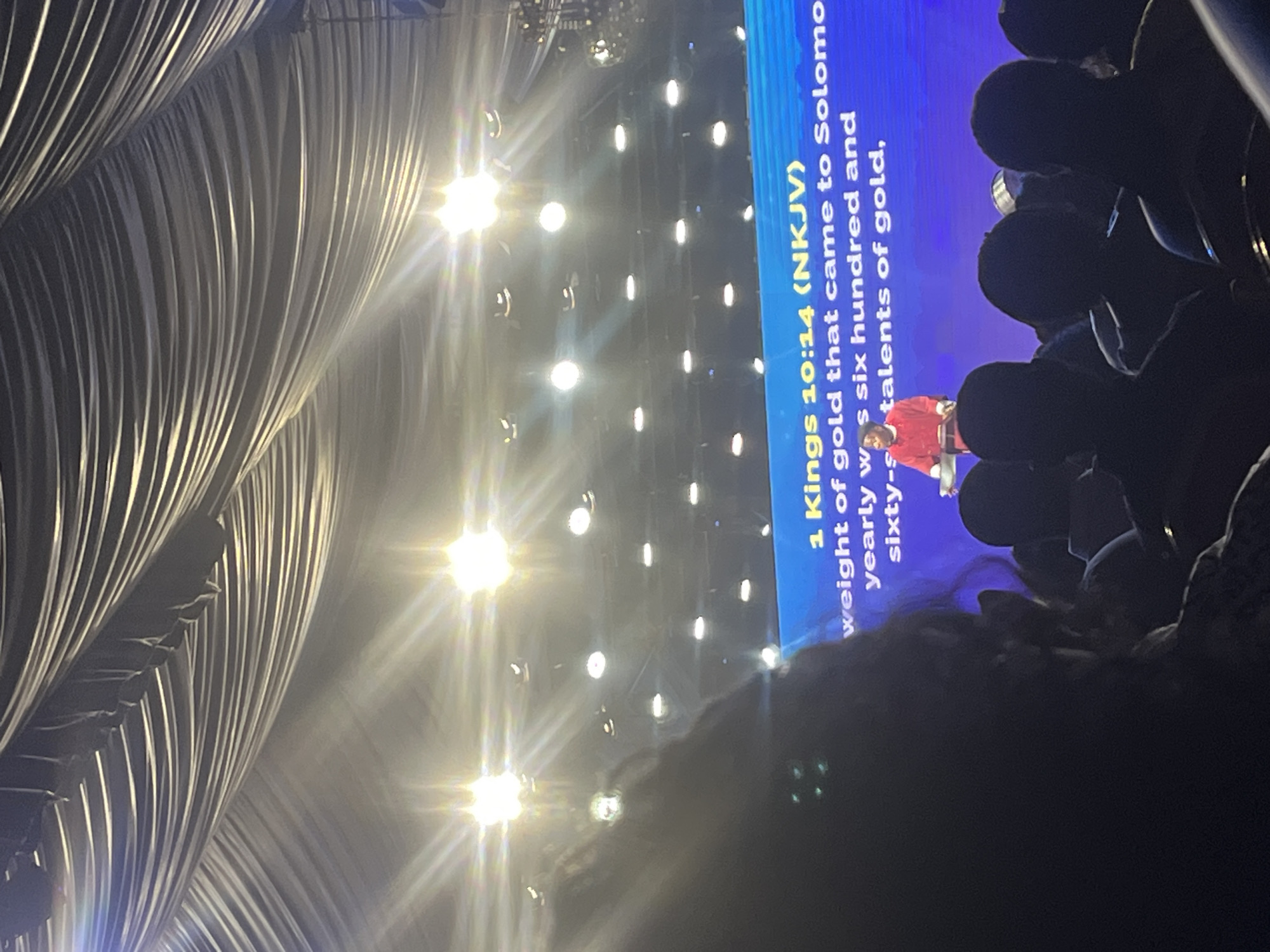 Here is a description of the image:
The image depicts a scene from an event likely related to religious practice, possibly in a large indoor space with stage lighting. In the foreground, there are silhouettes of people, likely audience members.
A screen displays text from the Bible, specifically 1 Kings 10:14 (NKJV), which discusses the weight of gold that came to Solomon yearly. A person, possibly a speaker or performer, is visible on the screen, wearing a red outfit.
The background features a complex structure of draped fabric, possibly part of the stage design, and a series of lights.