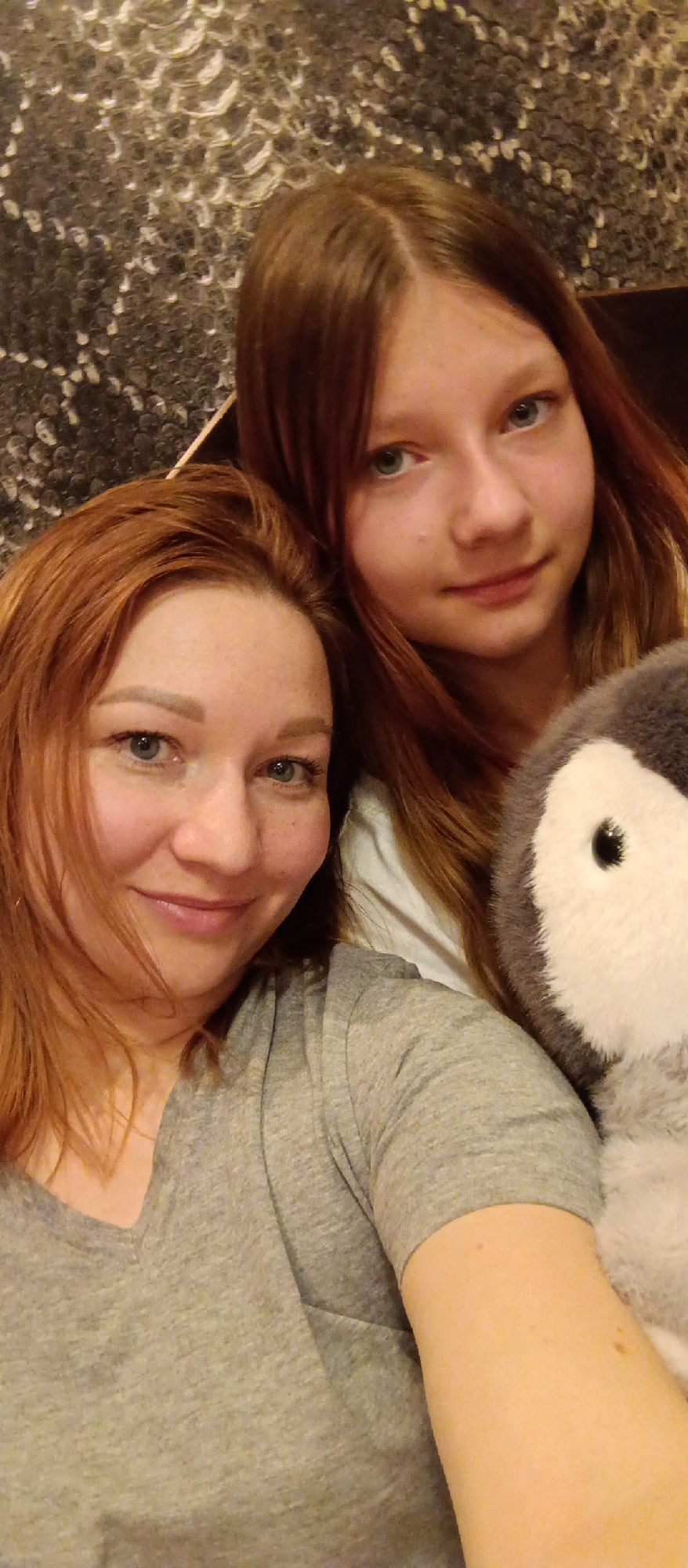 A close-up, eye-level portrait shows two females and a plush toy in an indoor setting. In the foreground on the left, an adult woman with reddish-brown hair and blue-green eyes smiles gently towards the viewer. She has a light complexion with visible freckles and is wearing a grey V-neck t-shirt. Behind her and slightly to the right is a younger female, appearing to be a teenager or pre-teen, with long reddish-brown hair parted in the middle and blue-green eyes. She also looks towards the camera with a subtle, soft smile and is wearing a light-colored top.
On the far right, next to the younger female, a grey and white plush toy resembling a penguin is partially visible. The two individuals are positioned closely, suggesting a familial or close relationship. The background features a dark, textured wall with a distinctive pattern of lighter, possibly reflective, raised circular or abstract shapes. The overall lighting is soft and appears to be artificial, contributing to a warm and intimate atmosphere. No specific location cues other than "indoors" are discernible, and there is no visible text. Both subjects convey a sense of calm and contentment through their gentle expressions.
