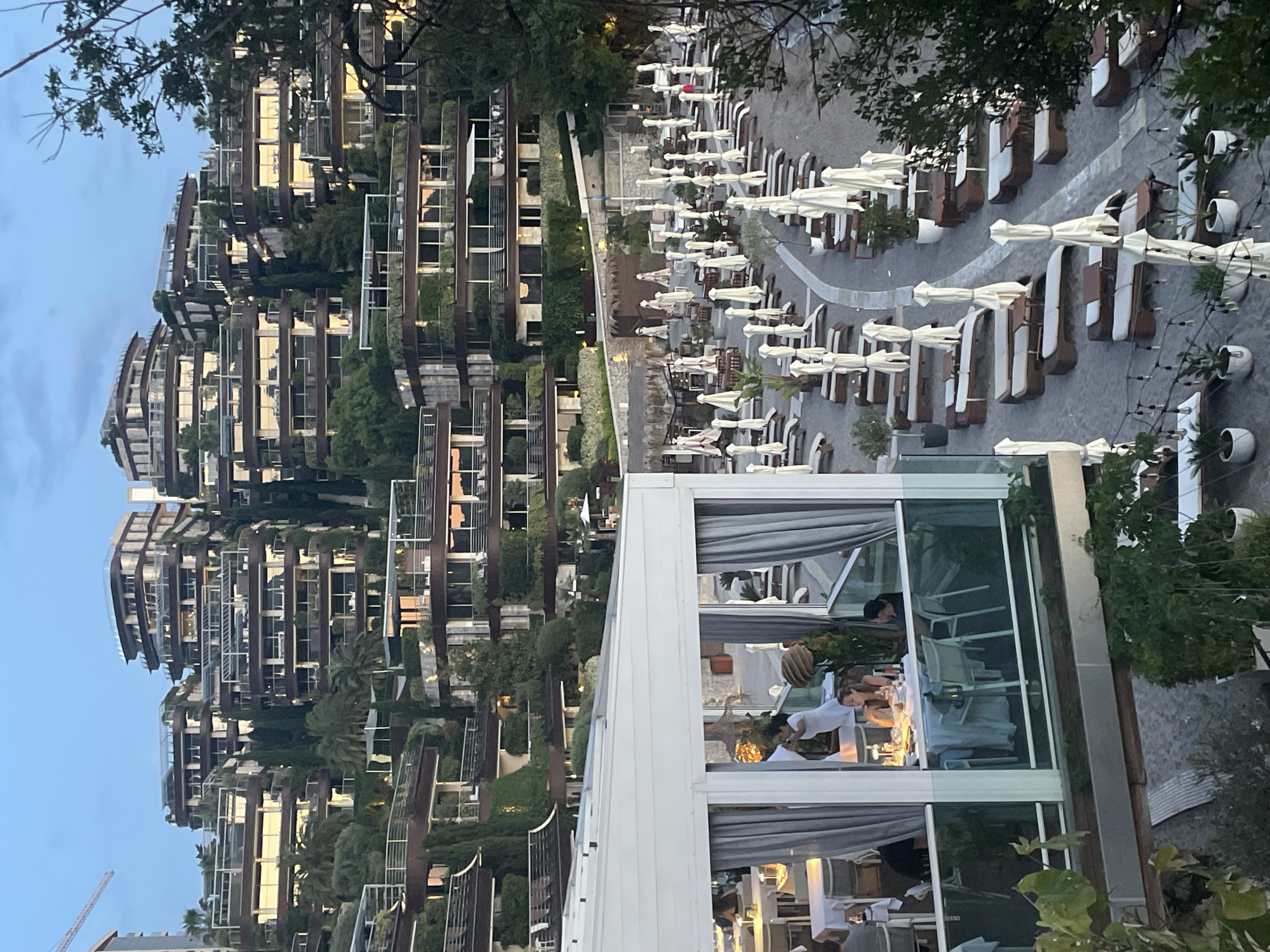 The image shows a view in the city of Budva, Montenegro. In the foreground is an outdoor dining area with a white-framed glass enclosure. People are seated at tables inside the enclosure. Beyond the dining area, there's a patio with numerous lounge chairs and white umbrellas. In the background, there's a uniquely designed residential building with layered balconies and greenery. The sky is dusky, suggesting either early morning or late evening.